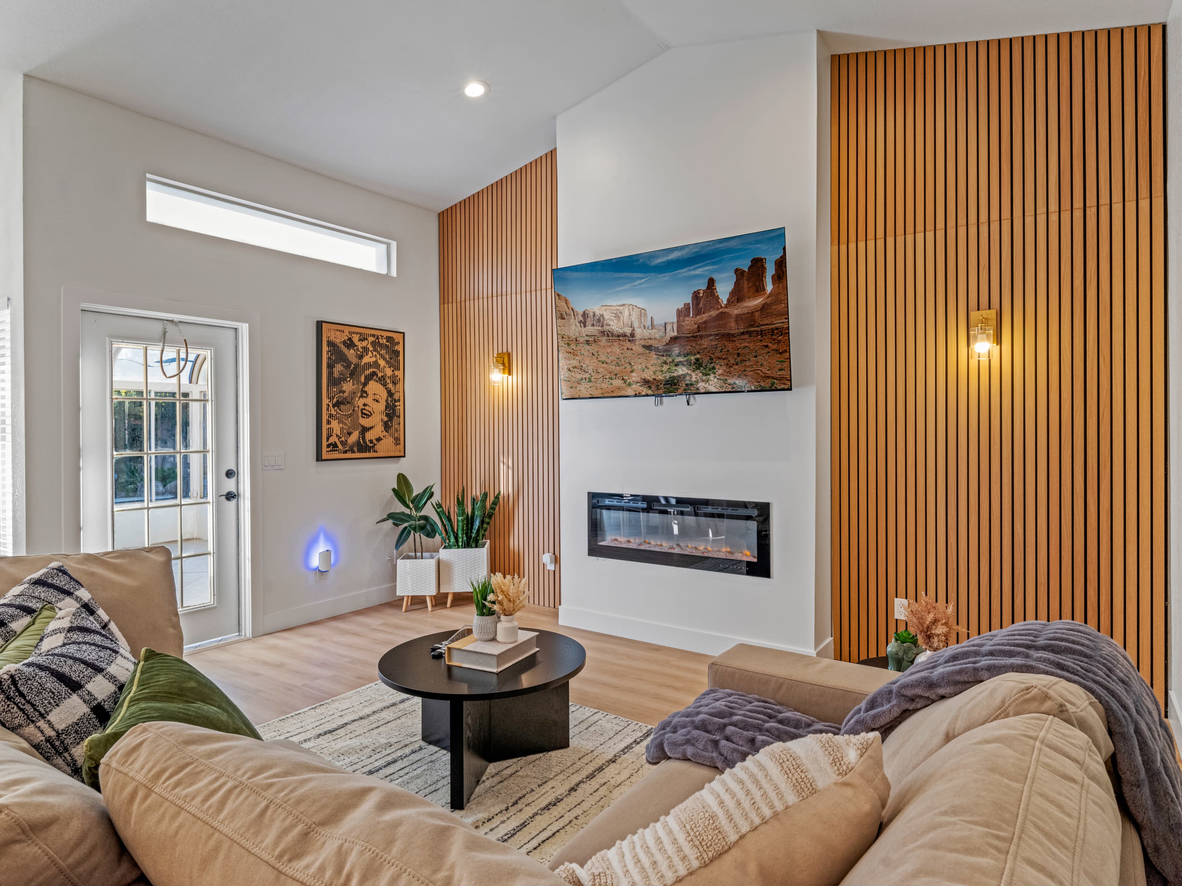 The living room features a mounted television above a modern fireplace, with wood paneling, a round coffee table, and a large window allowing natural light.