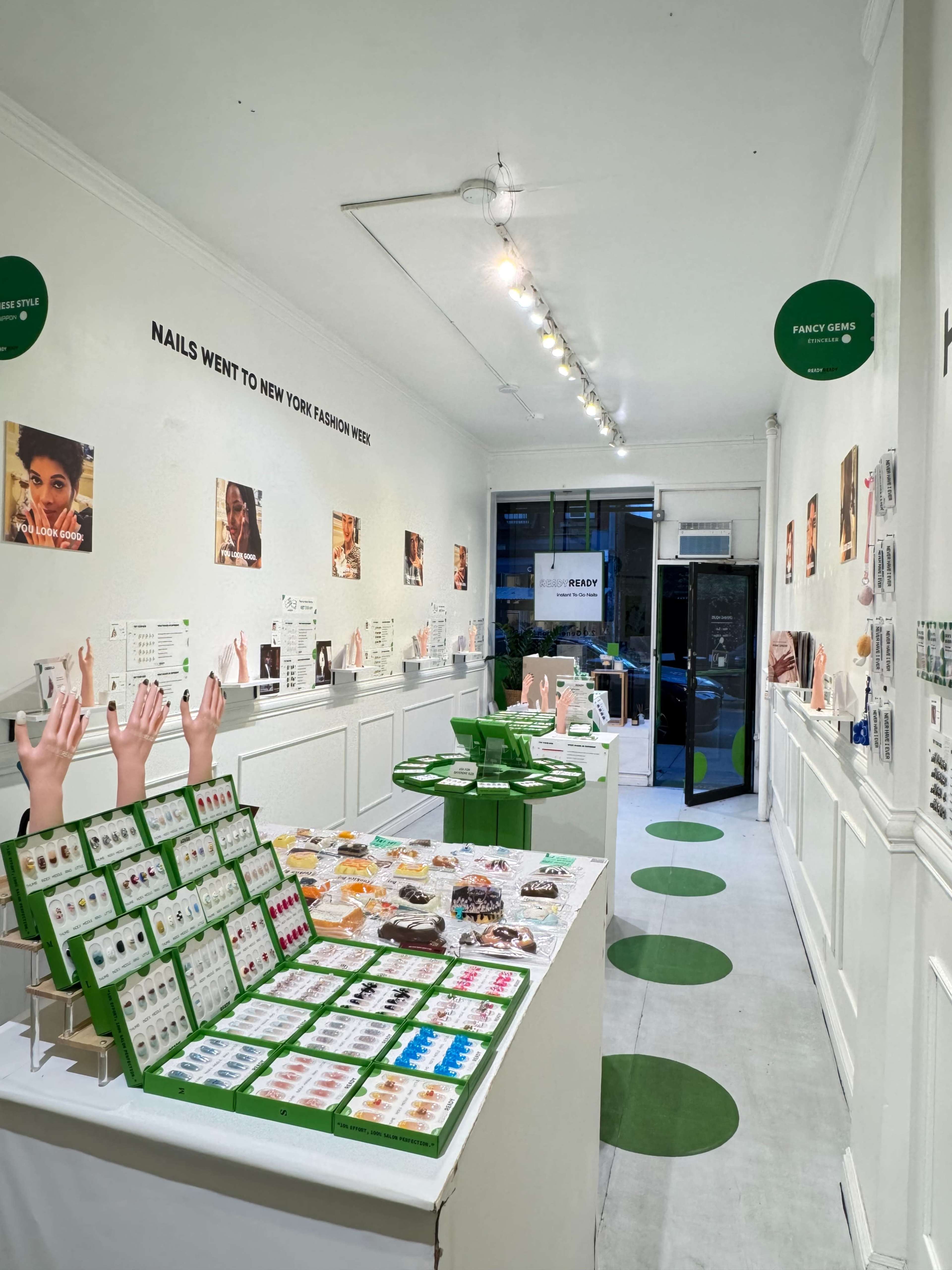 The image shows an interior view of a retail space featuring a variety of nail products arranged on tables, with posters and promotional materials displayed on the walls.