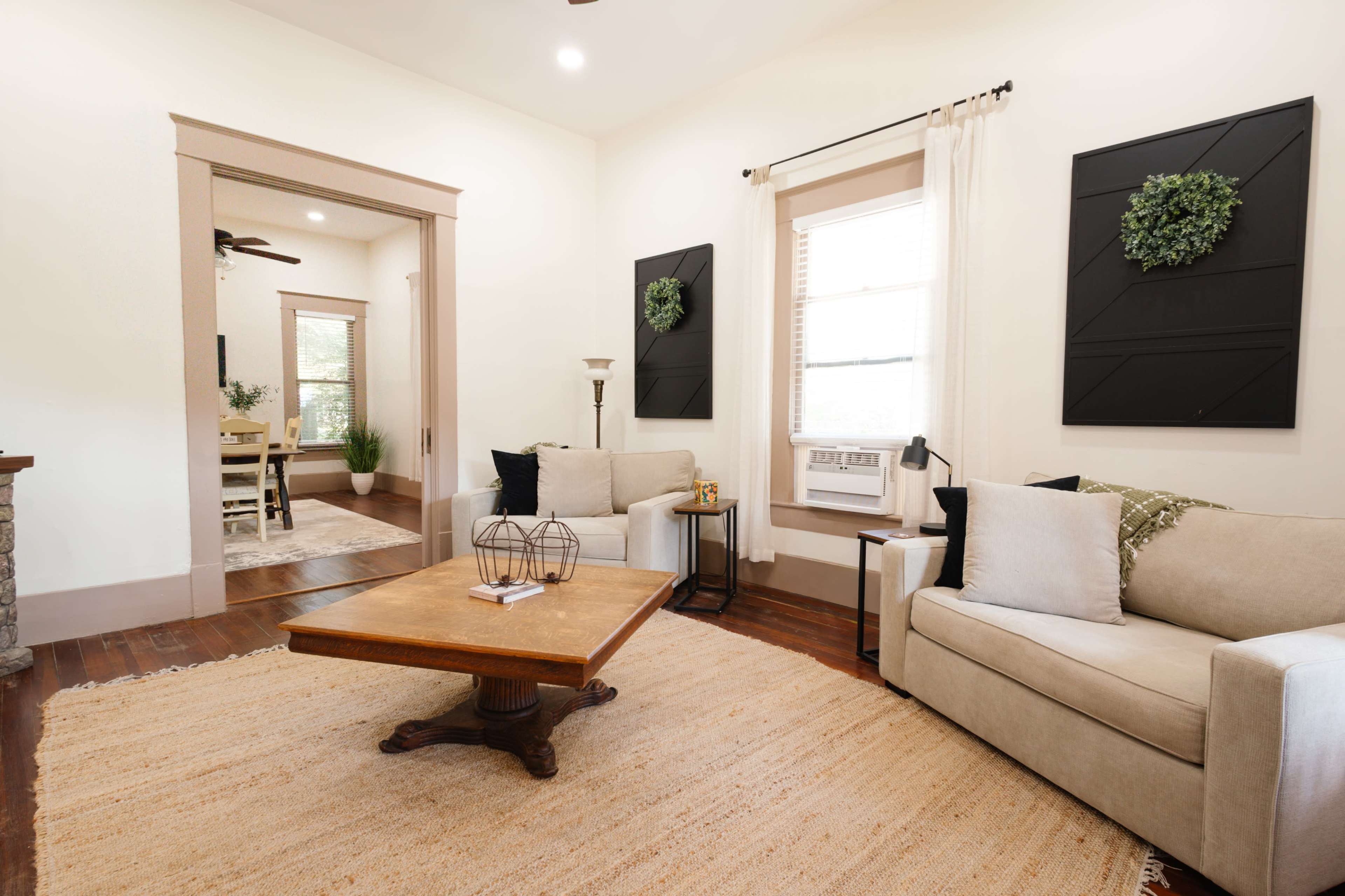 A cozy living room features a beige sofa, a wooden coffee table, and decorative wall art, with an adjacent room visible through an open archway.