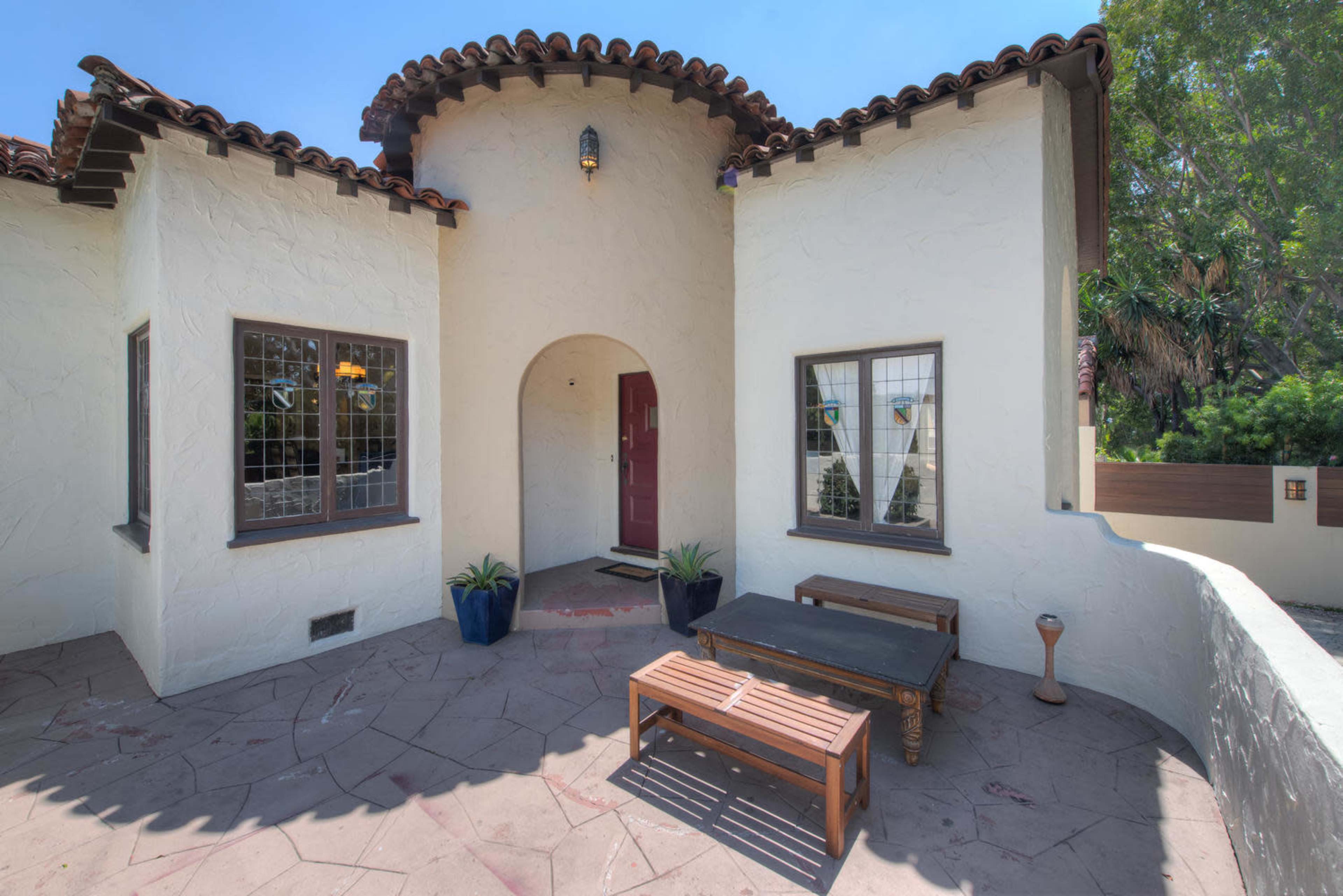 The entrance of a Mediterranean-style house featuring a rounded archway, a red door, and a patio with a wooden bench and potted plants.