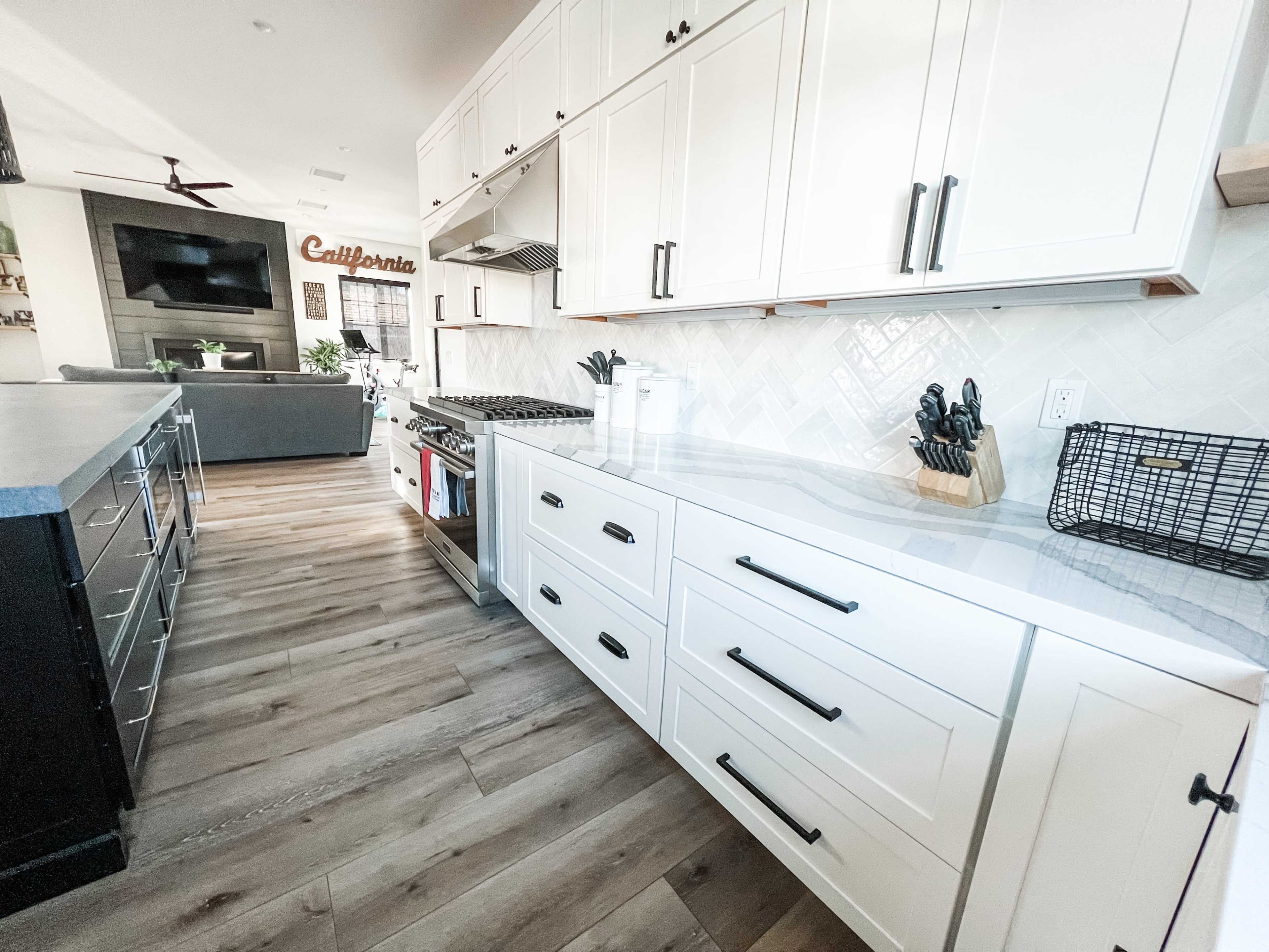A modern kitchen featuring white cabinets, a gray countertop, and stainless steel appliances, with distinct wood flooring and a living area visible in the background.