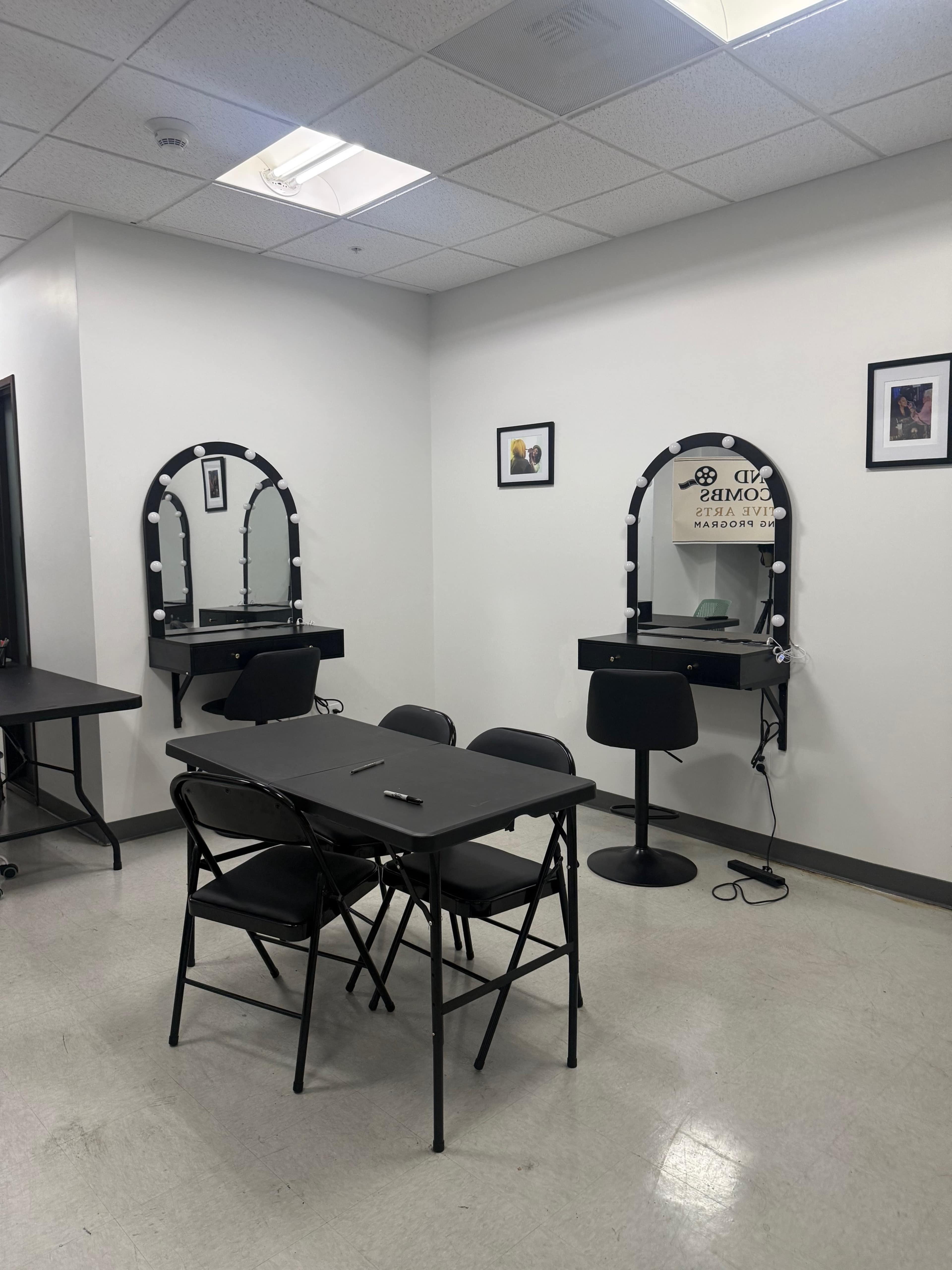 The image shows a small room with two illuminated mirrors on the walls, a black folding table with chairs, and a hairdryer on the floor.