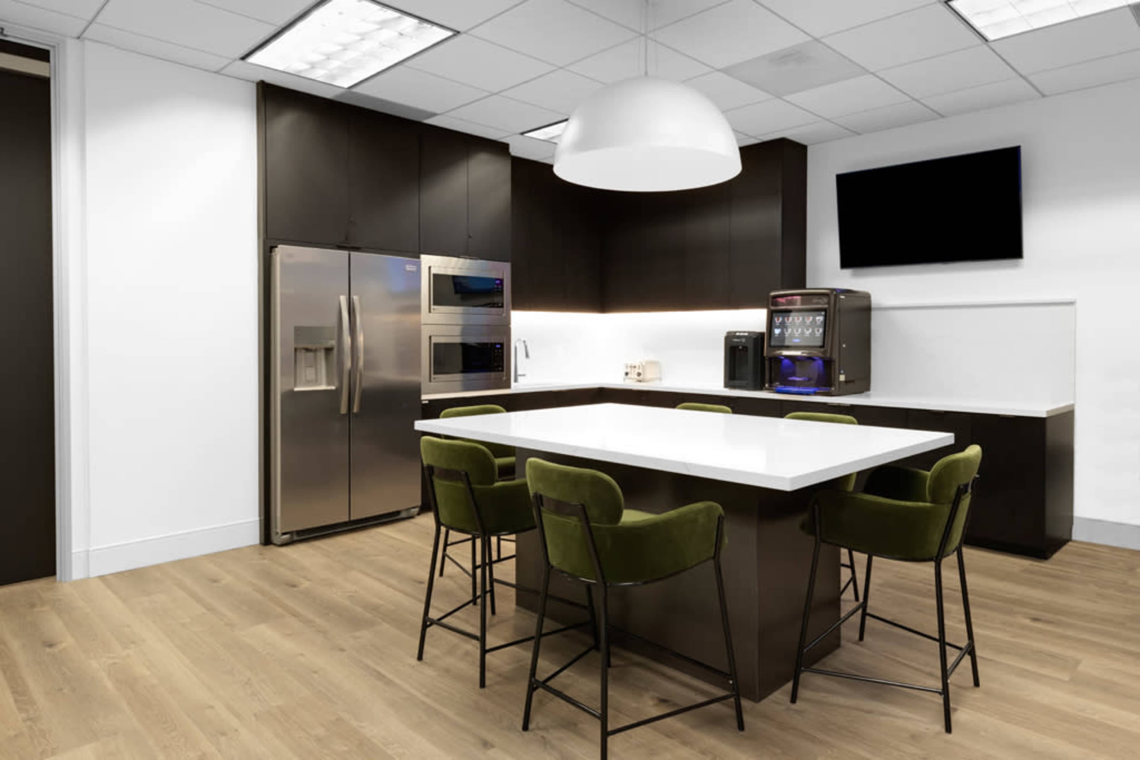 The image shows a modern office kitchen with a white island countertop, dark cabinetry, stainless steel appliances, and green bar stools.