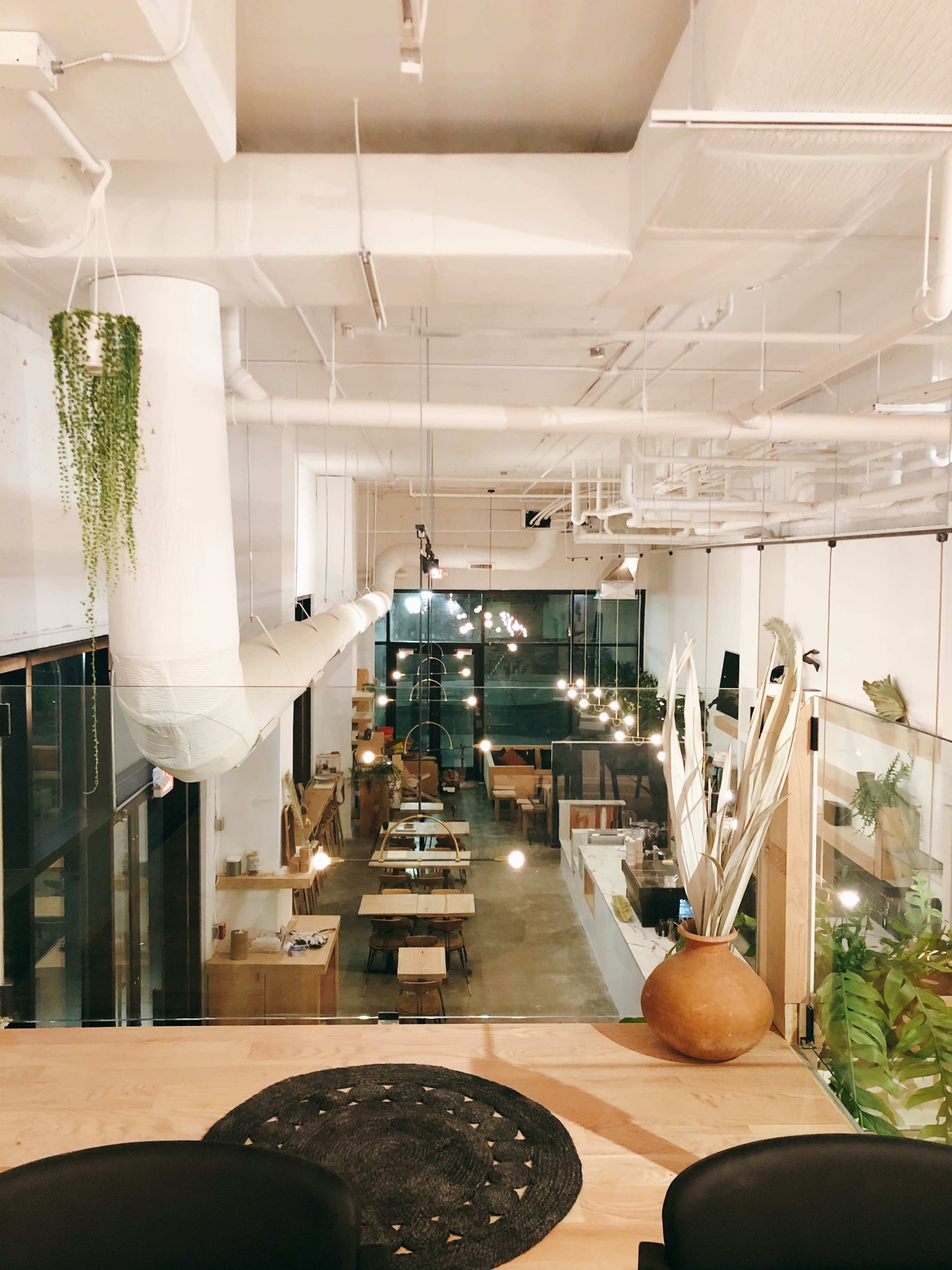 The image shows a spacious, modern workspace with wooden tables, hanging lights, and a clear view of a kitchen area, surrounded by plants and glass walls.