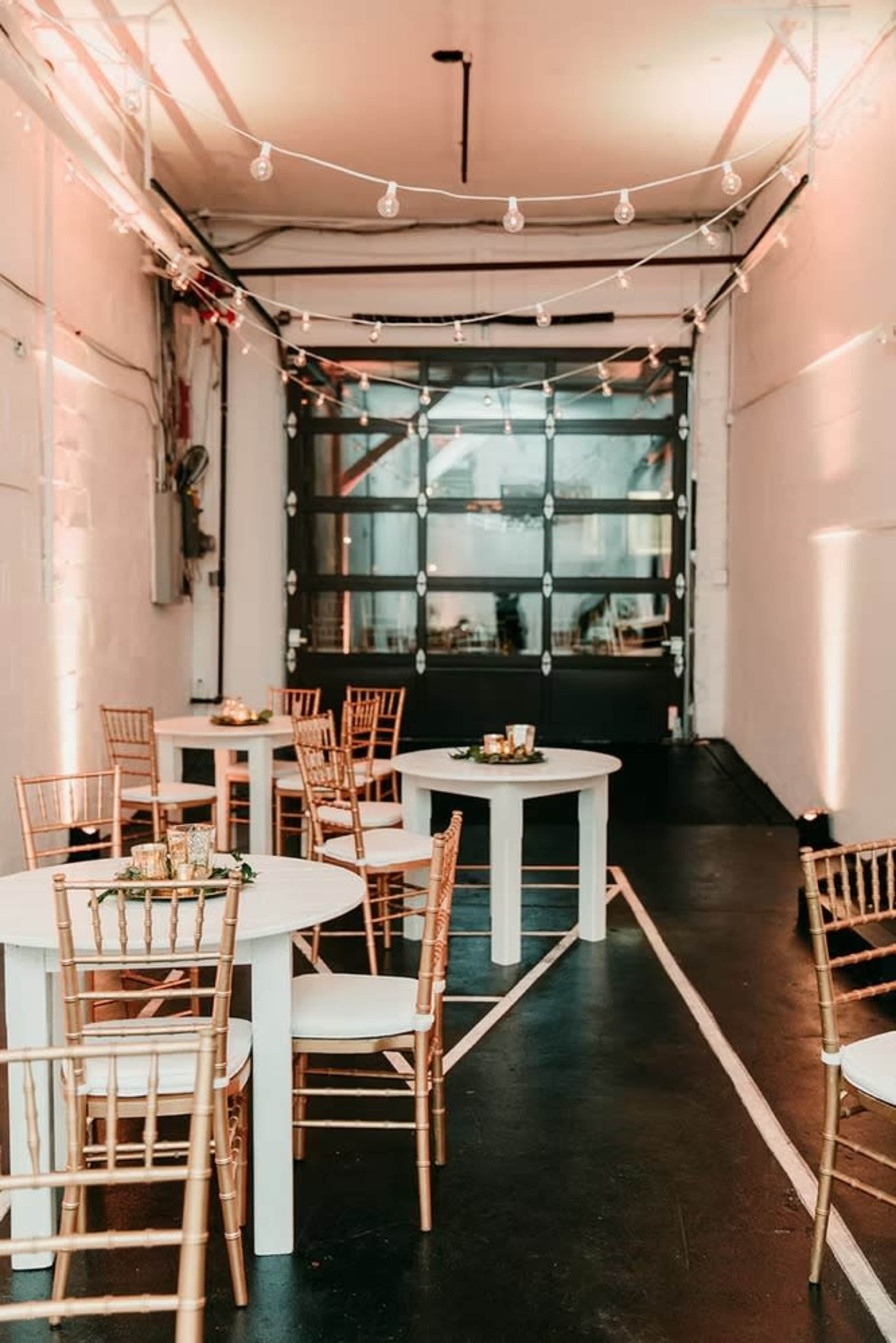 The image shows a modern event space featuring round white tables and gold chiavari chairs, illuminated by string lights along the ceiling.