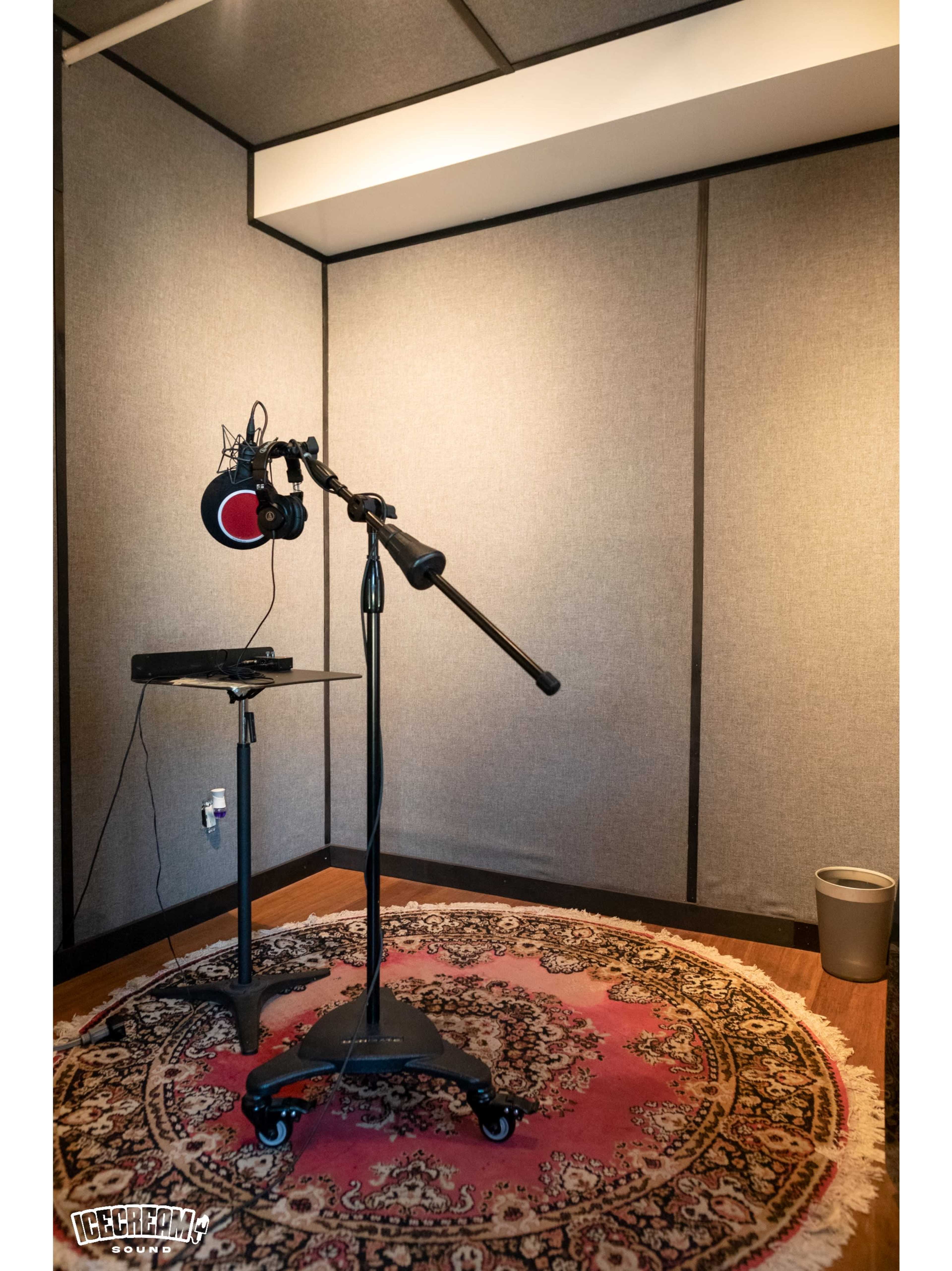 The image shows a soundproof recording booth with a microphone on a stand, a small table, and a decorative rug on the floor.