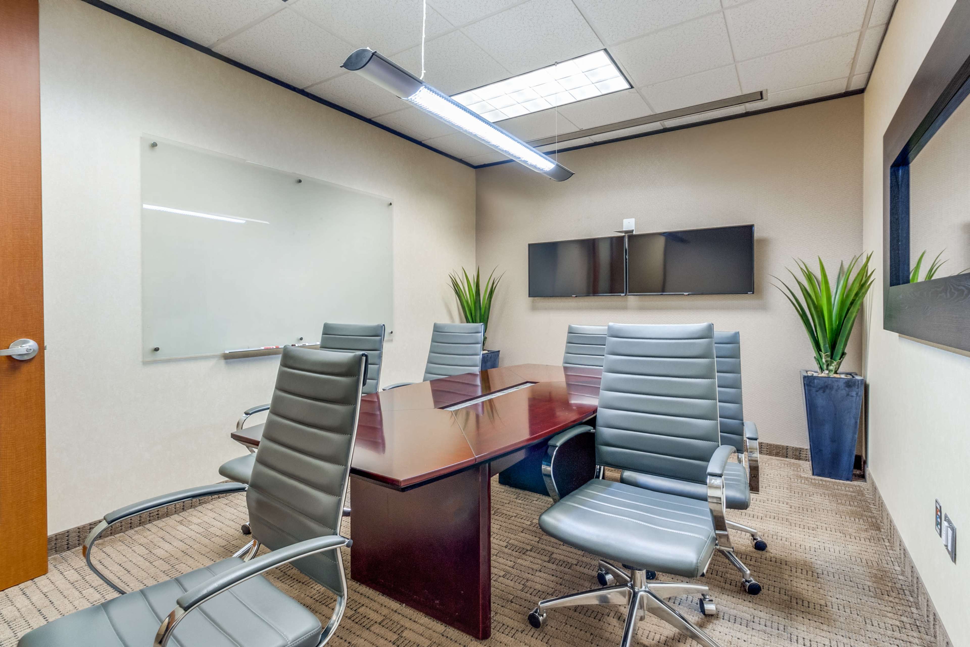 A modern conference room with a wooden table, six metal chairs, a whiteboard, and two wall-mounted screens.