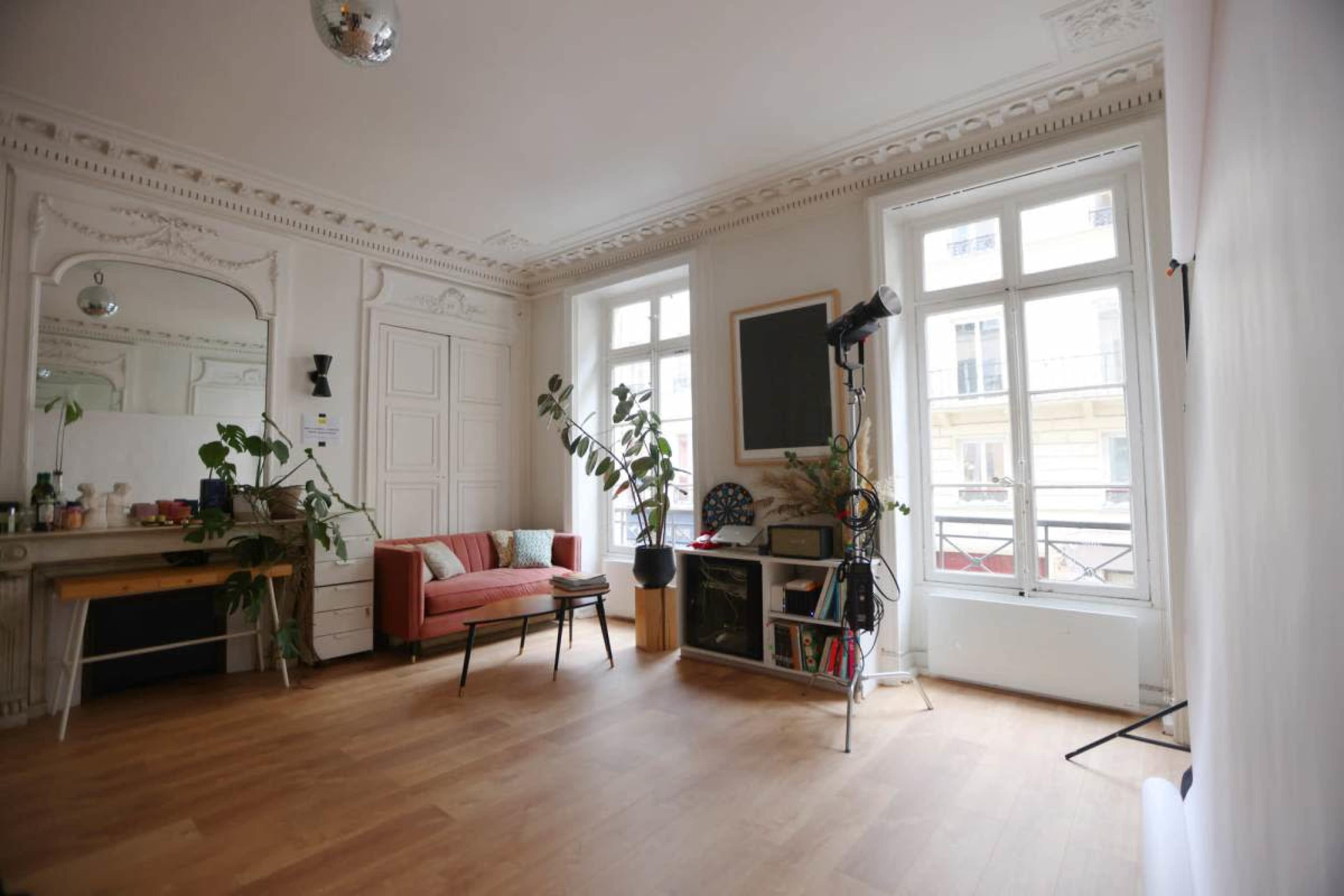 The room features a stylish interior with a pink sofa, a large mirror, houseplants, and a window providing natural light.