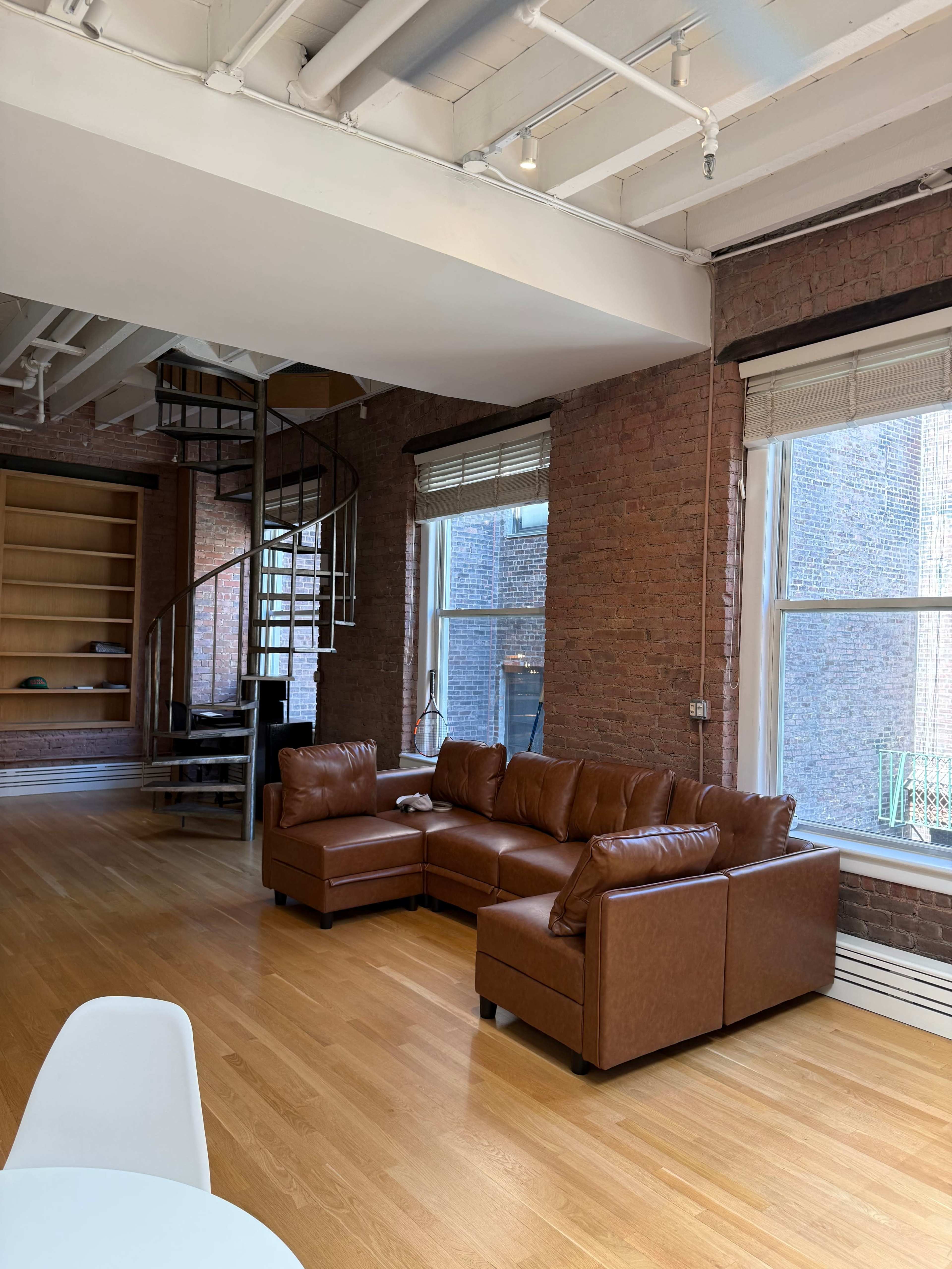 The image shows a spacious room with a brown leather sectional sofa, large windows, exposed brick walls, and a spiral staircase leading to an upper level.