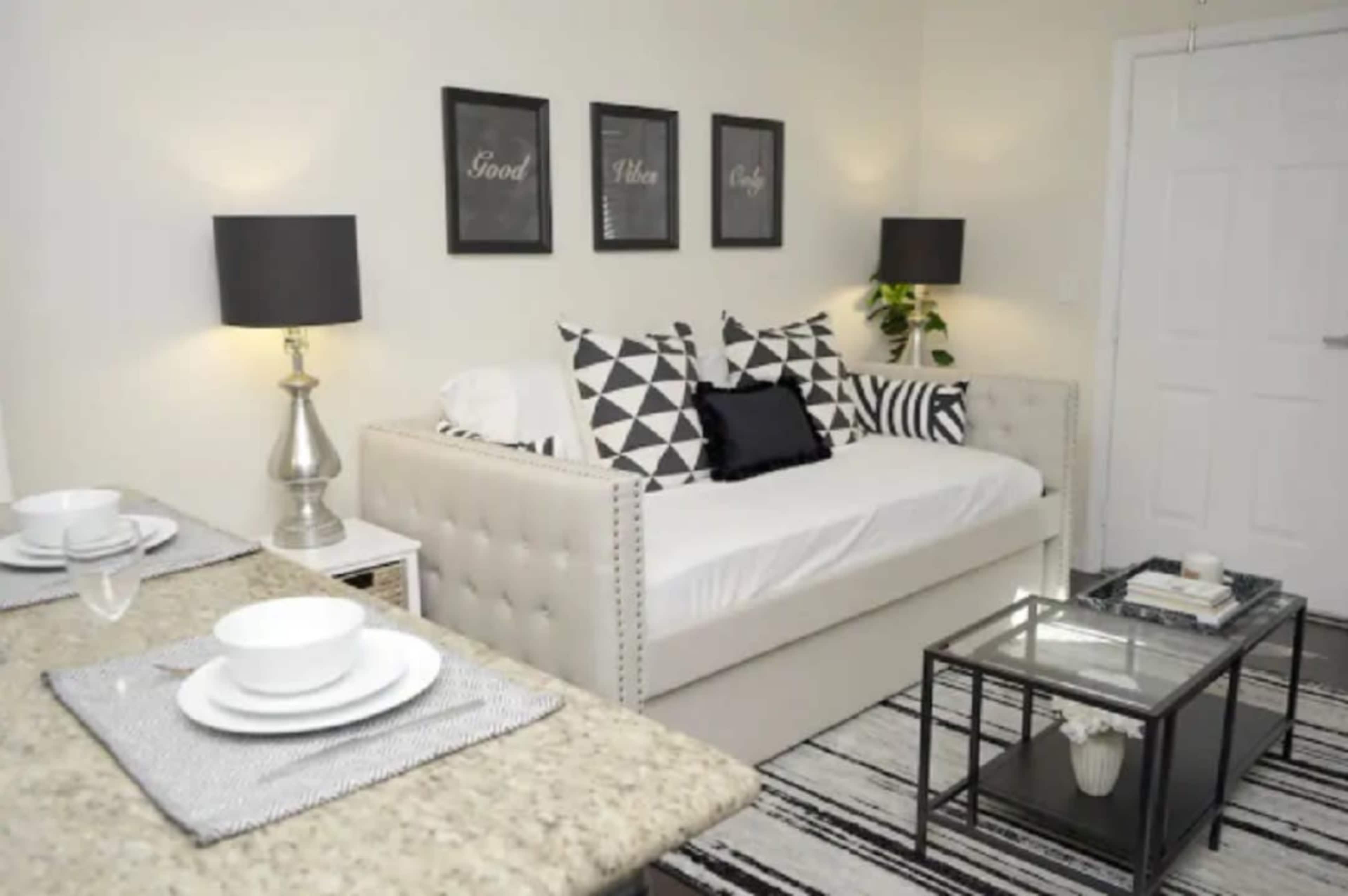 The image shows a modern living space featuring a daybed adorned with geometric-patterned cushions, a glass coffee table, and a dining area set with dishes on a granite countertop.