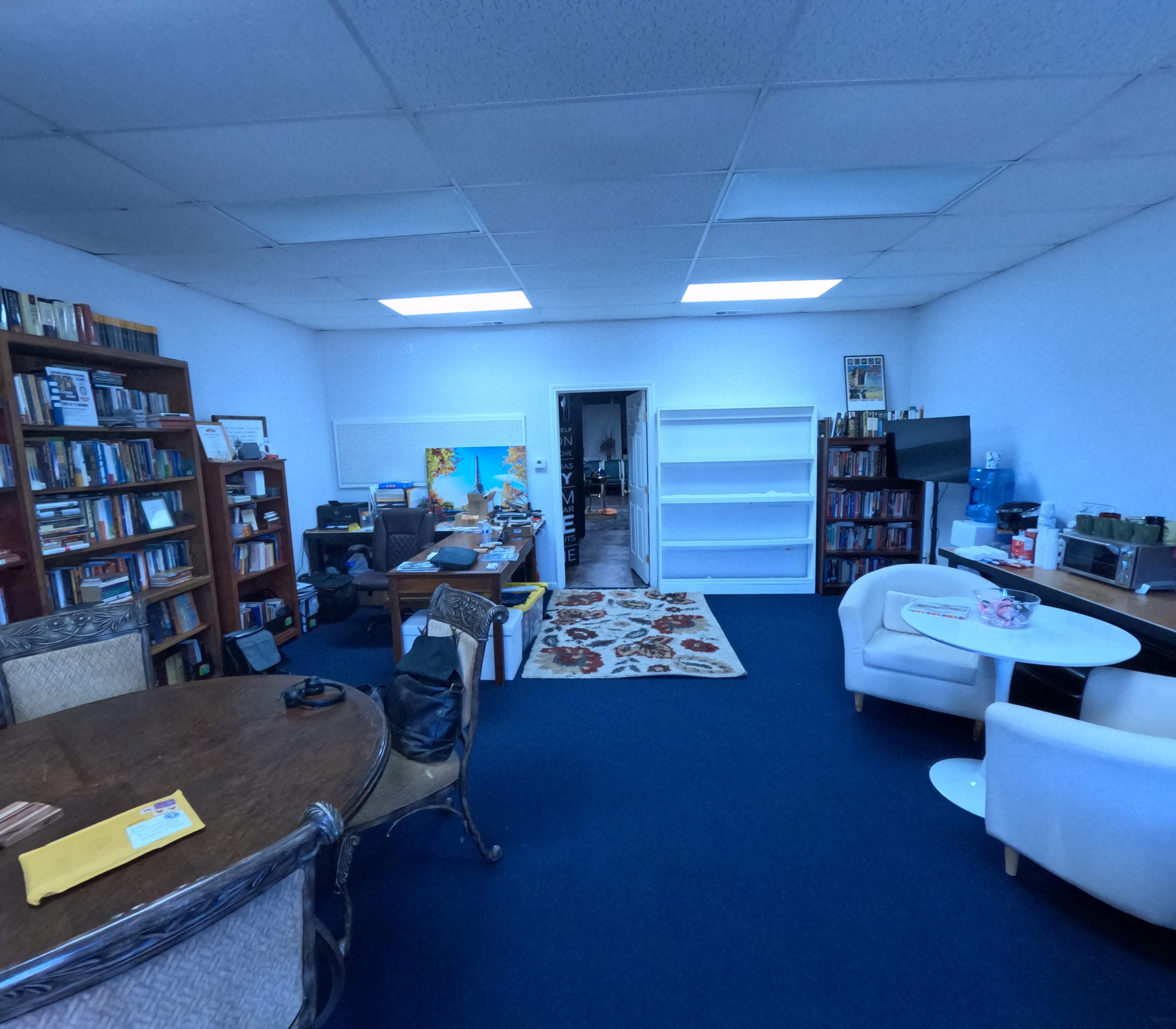 The image shows an office space with a large round wooden table, bookshelves filled with books, a couch, and a small dining table in a corner, along with a doorway leading to another room.