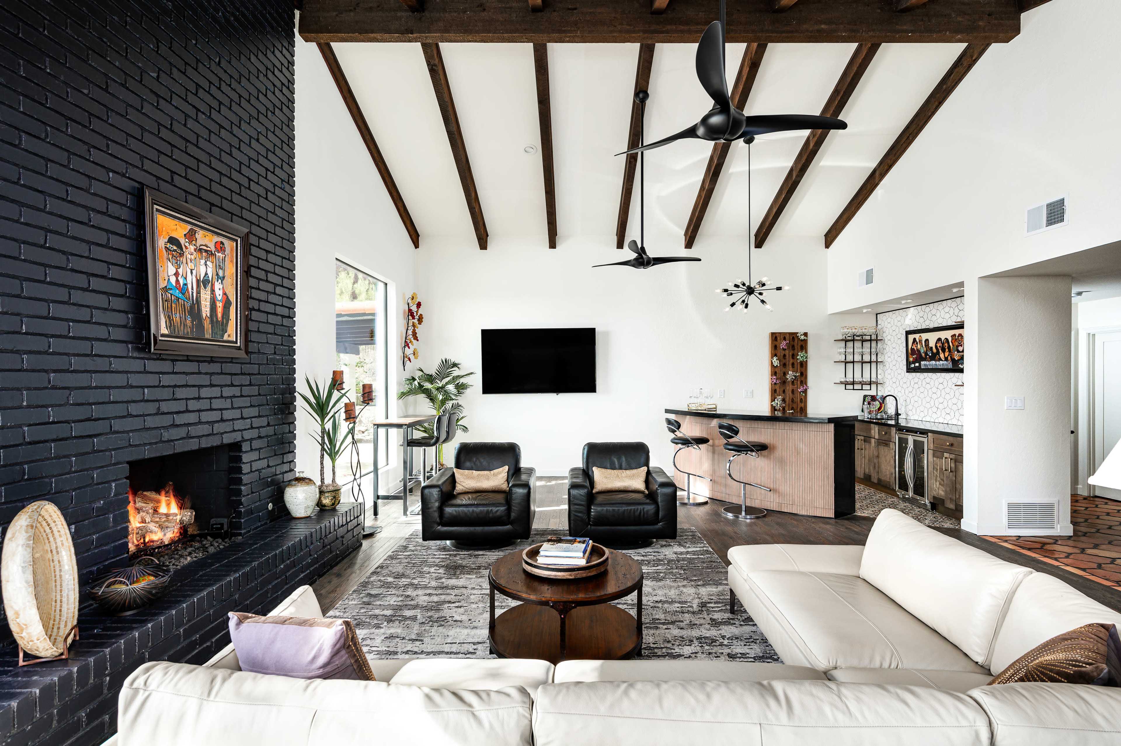A modern living room featuring a white sectional sofa, a round coffee table, a black brick fireplace, and a flat-screen TV mounted on the wall, with exposed wooden beams overhead.