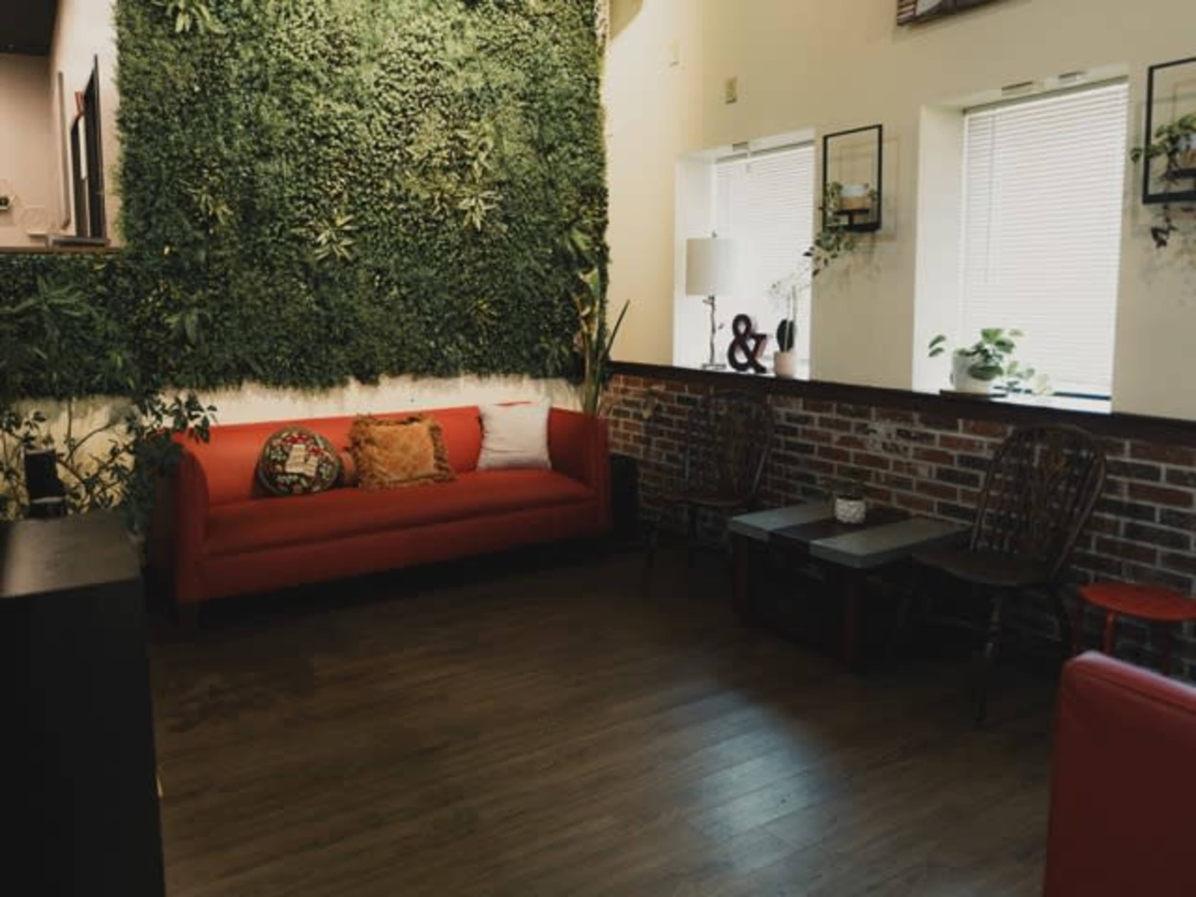 The image shows a cozy indoor space featuring a red sofa against a green plant wall, with wooden flooring and a brick accent wall.