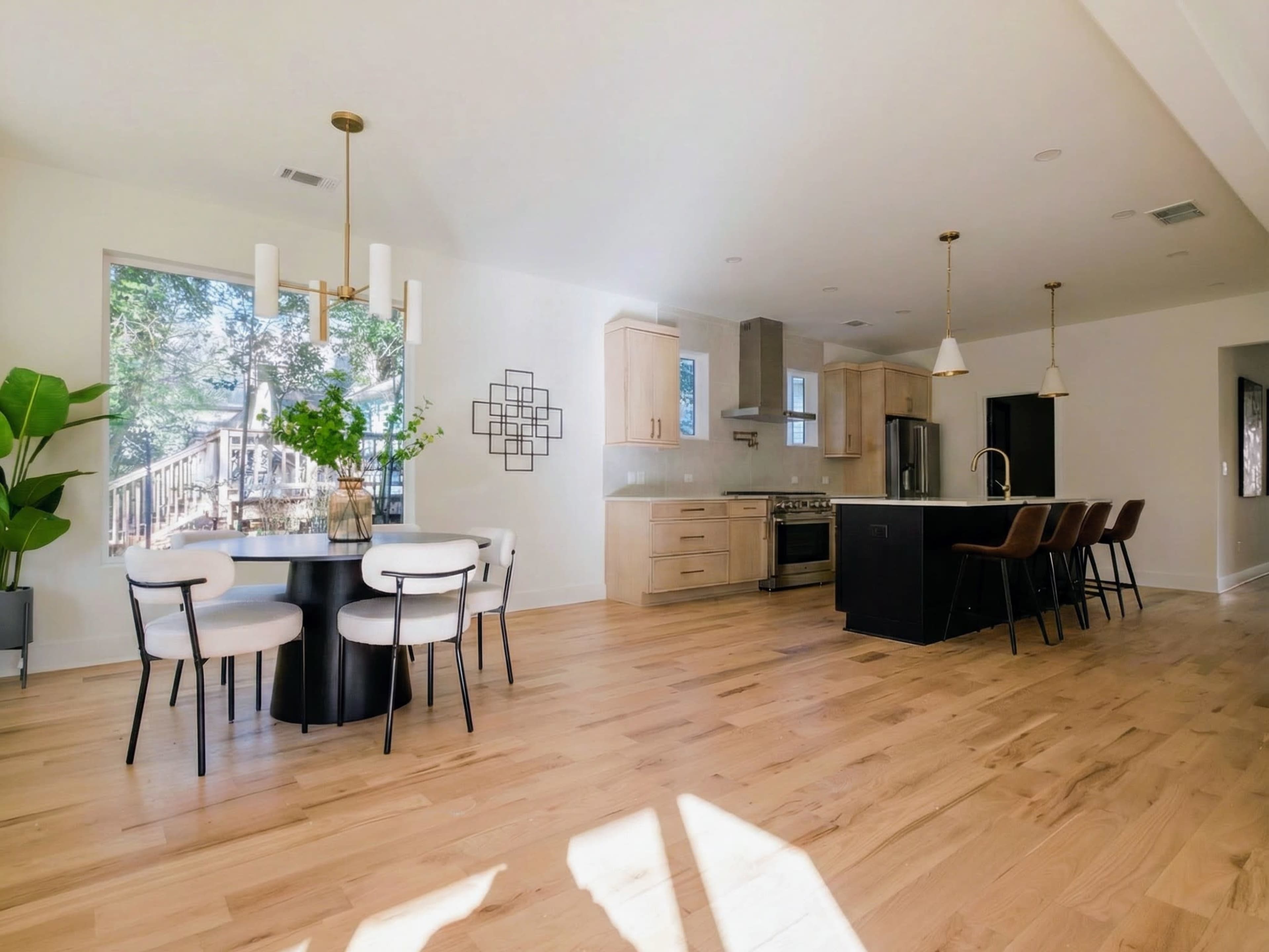 The image shows a modern kitchen and dining area featuring a round table with four chairs, an island with bar seating, and a large window that provides natural light and a view of an outdoor deck.