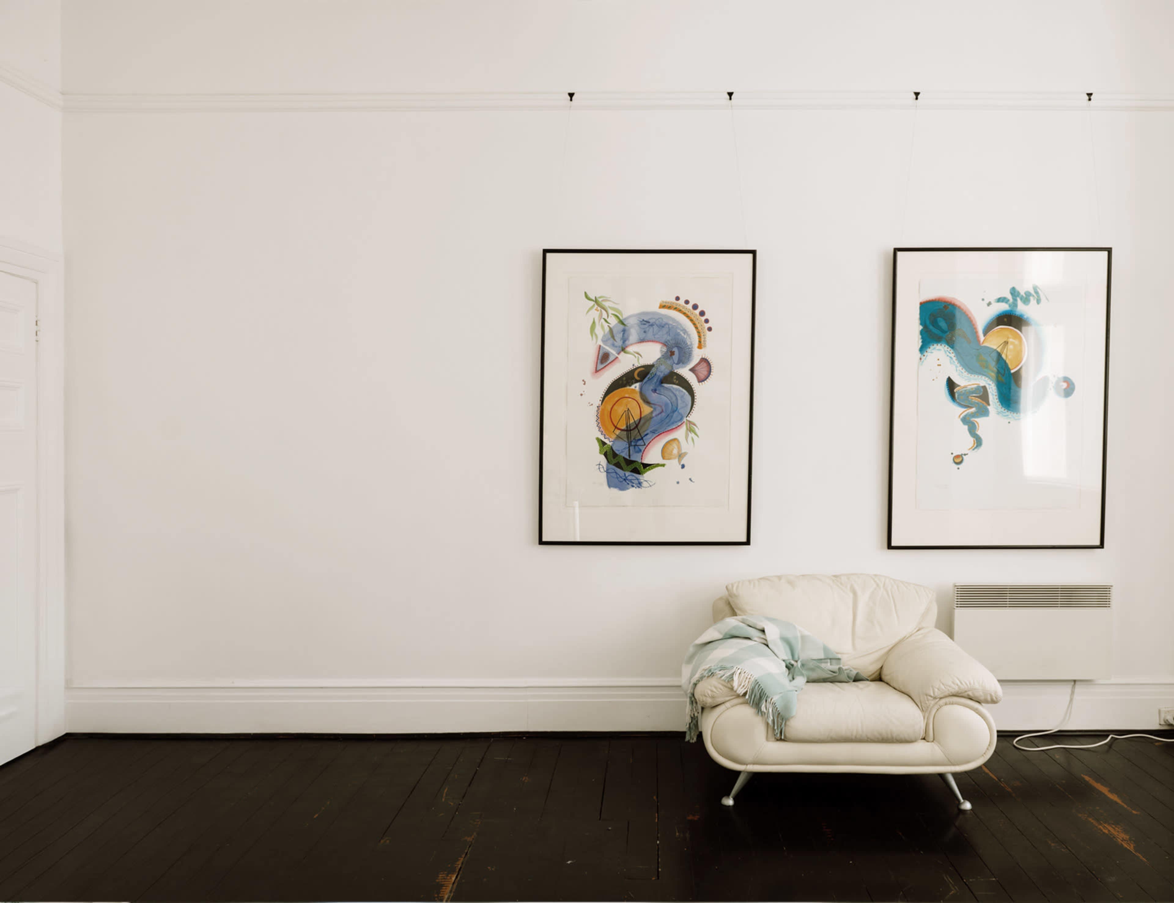 A minimalist room with two framed abstract paintings on a white wall and a white chair draped with a striped blanket.