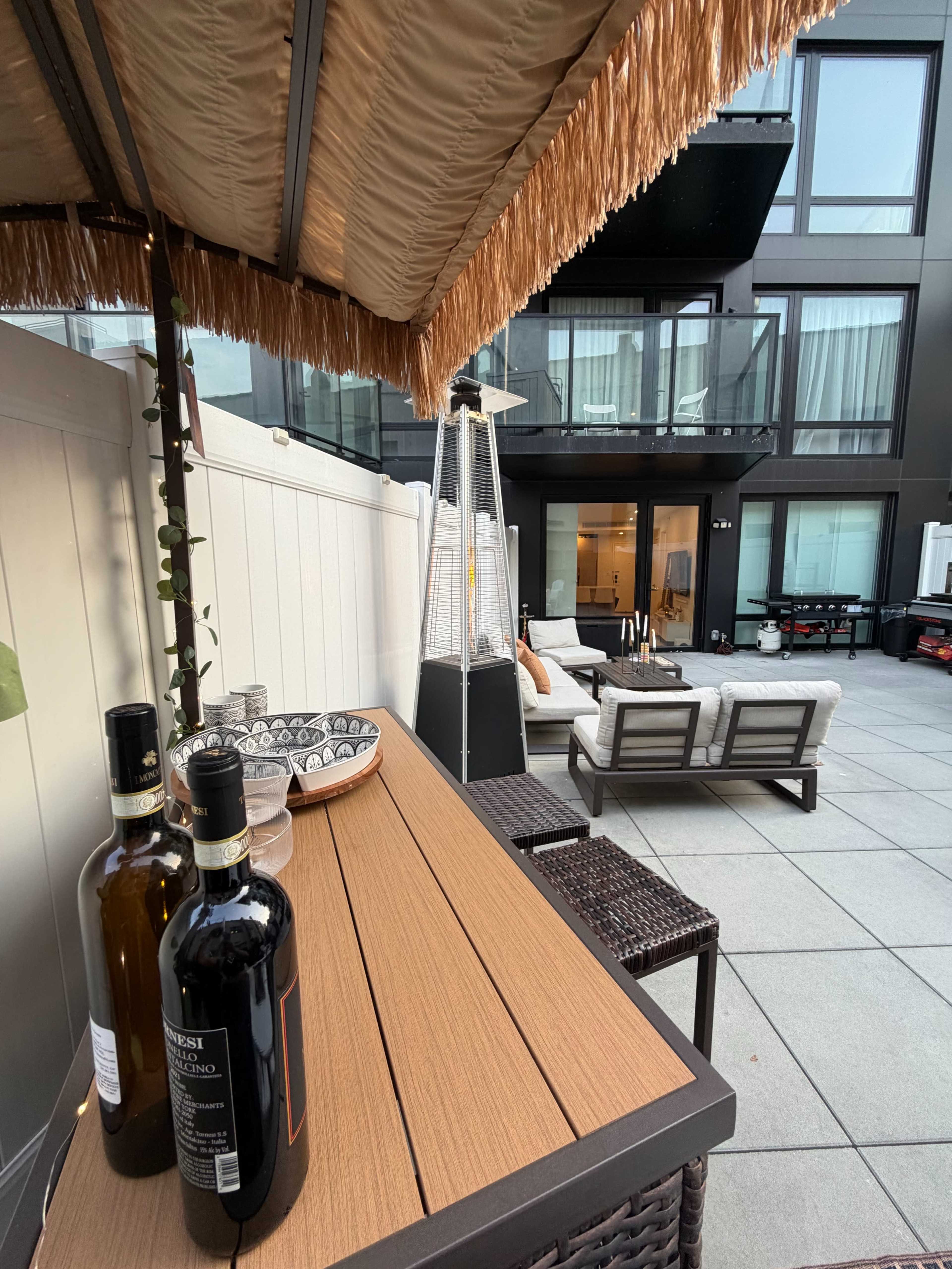 The image shows a modern outdoor patio area featuring a bar cart with bottles, seating arrangements, and a backdrop of a multi-story building.
