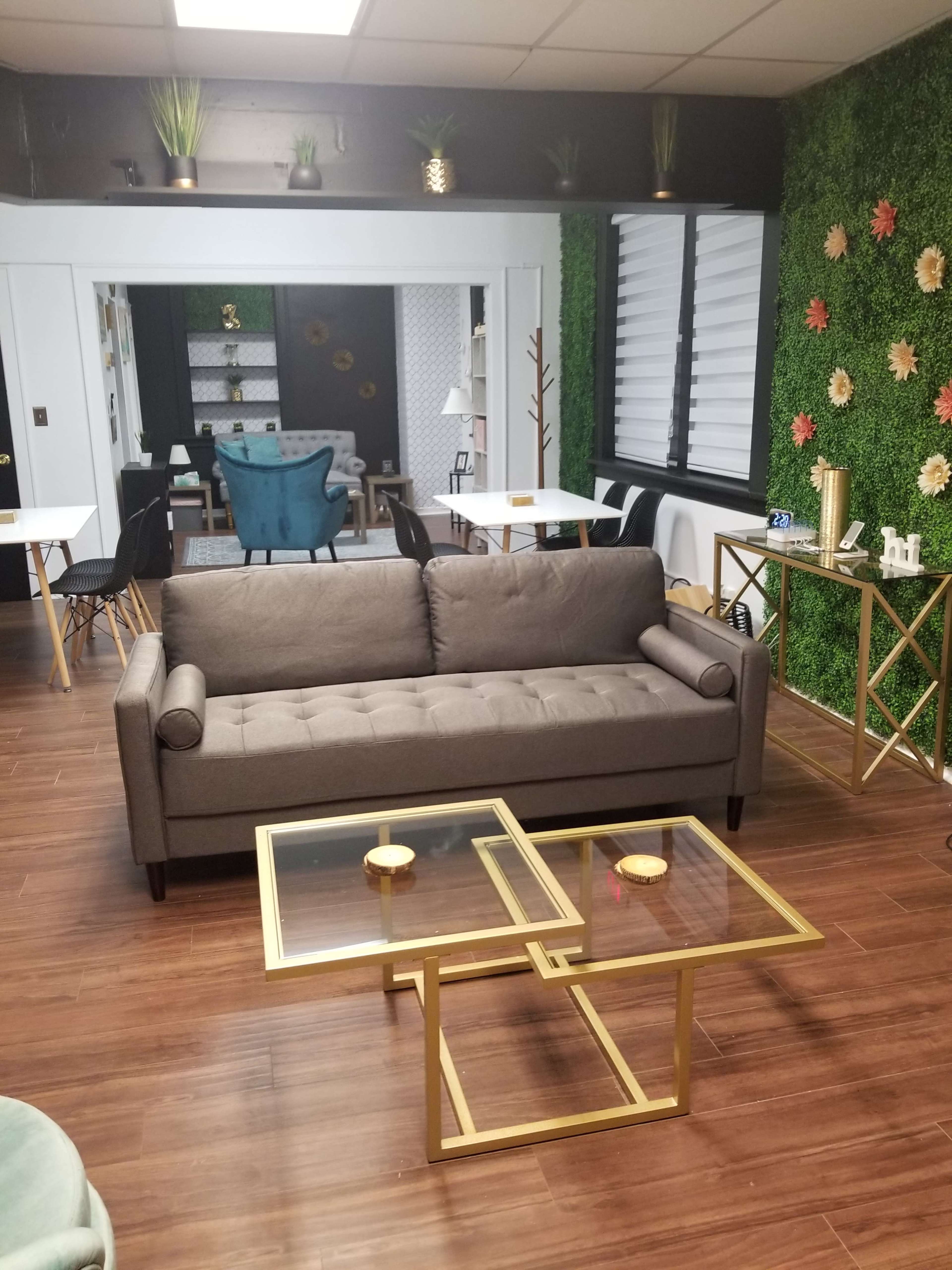A modern lounge area features a gray sofa, two glass coffee tables with gold frames, and greenery on the walls.