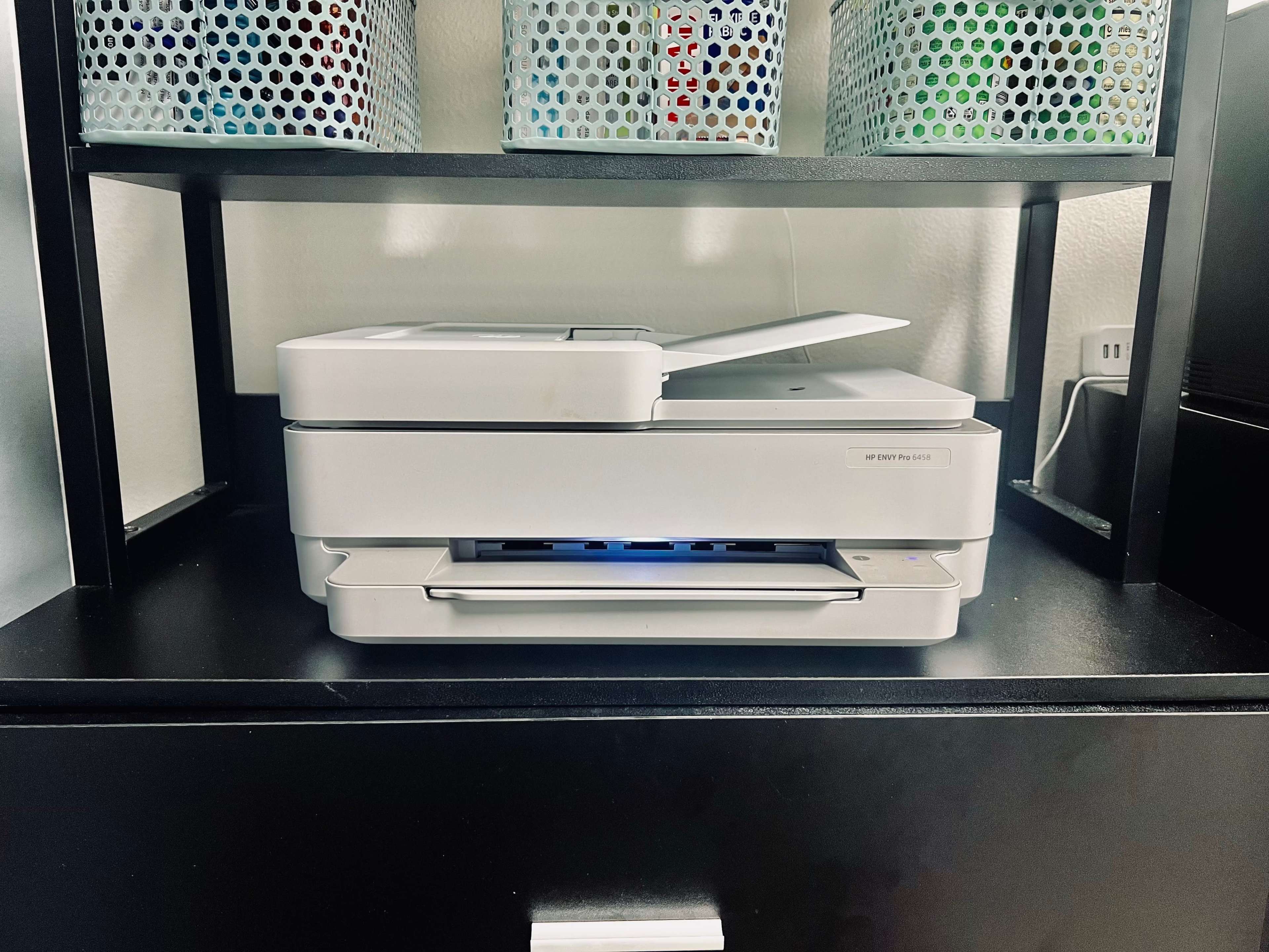 A white printer and scanner is placed on a black surface, with metallic baskets containing items above it.