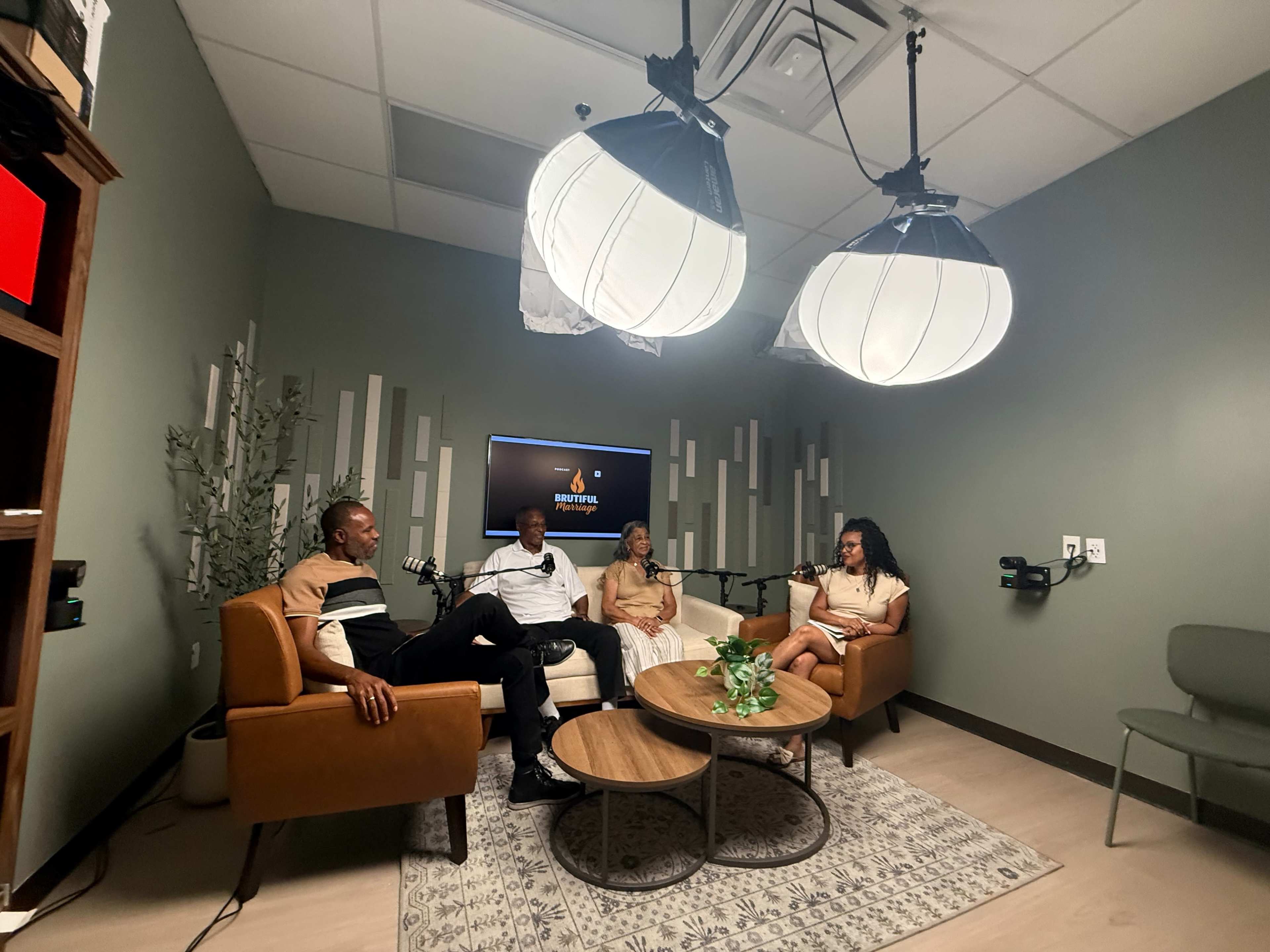 Four people sit in a podcast studio equipped with a round table, microphones, and large overhead lights.