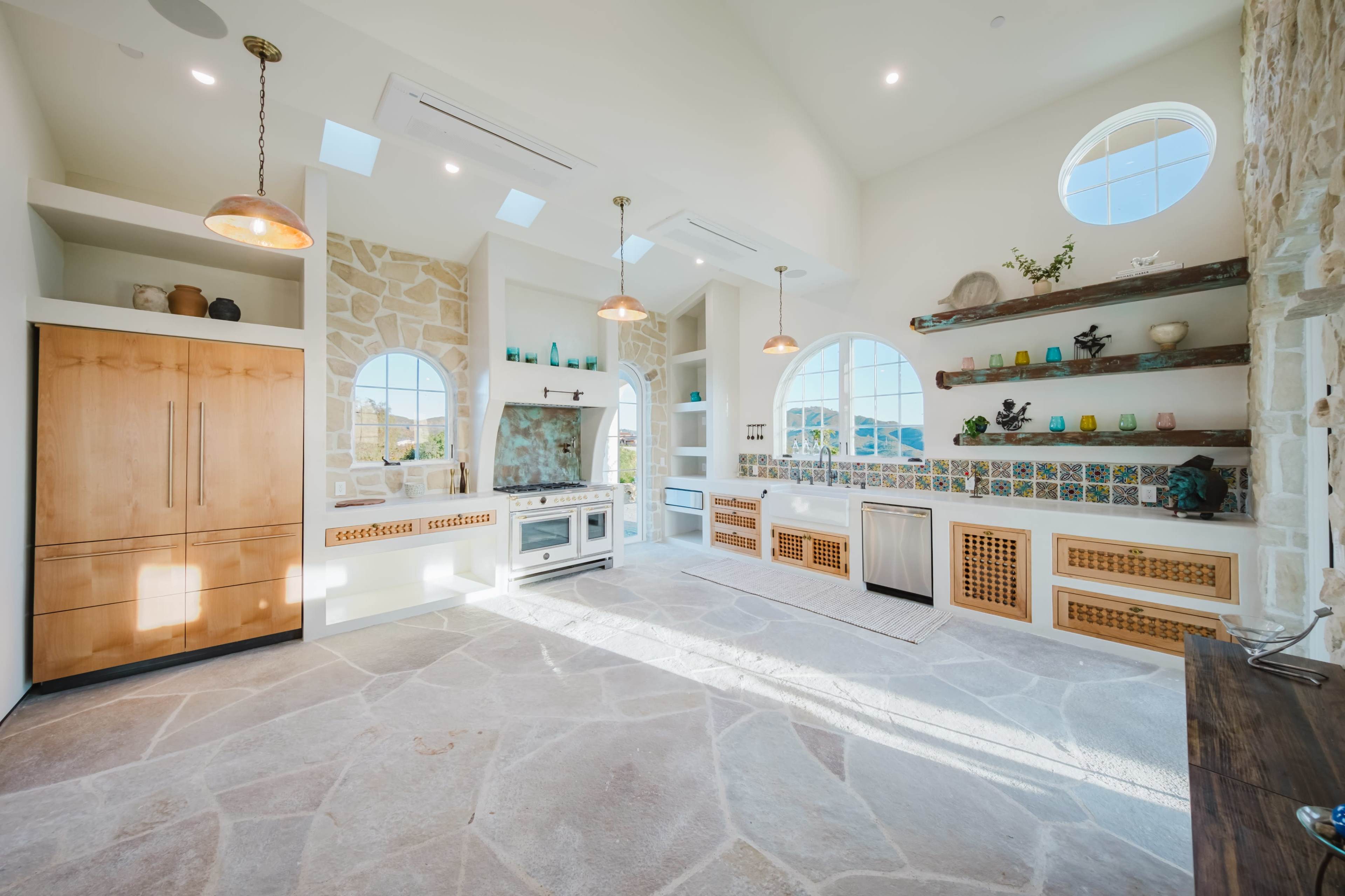 The image shows a bright, spacious kitchen with large windows, stone flooring, and decorative tile accents.