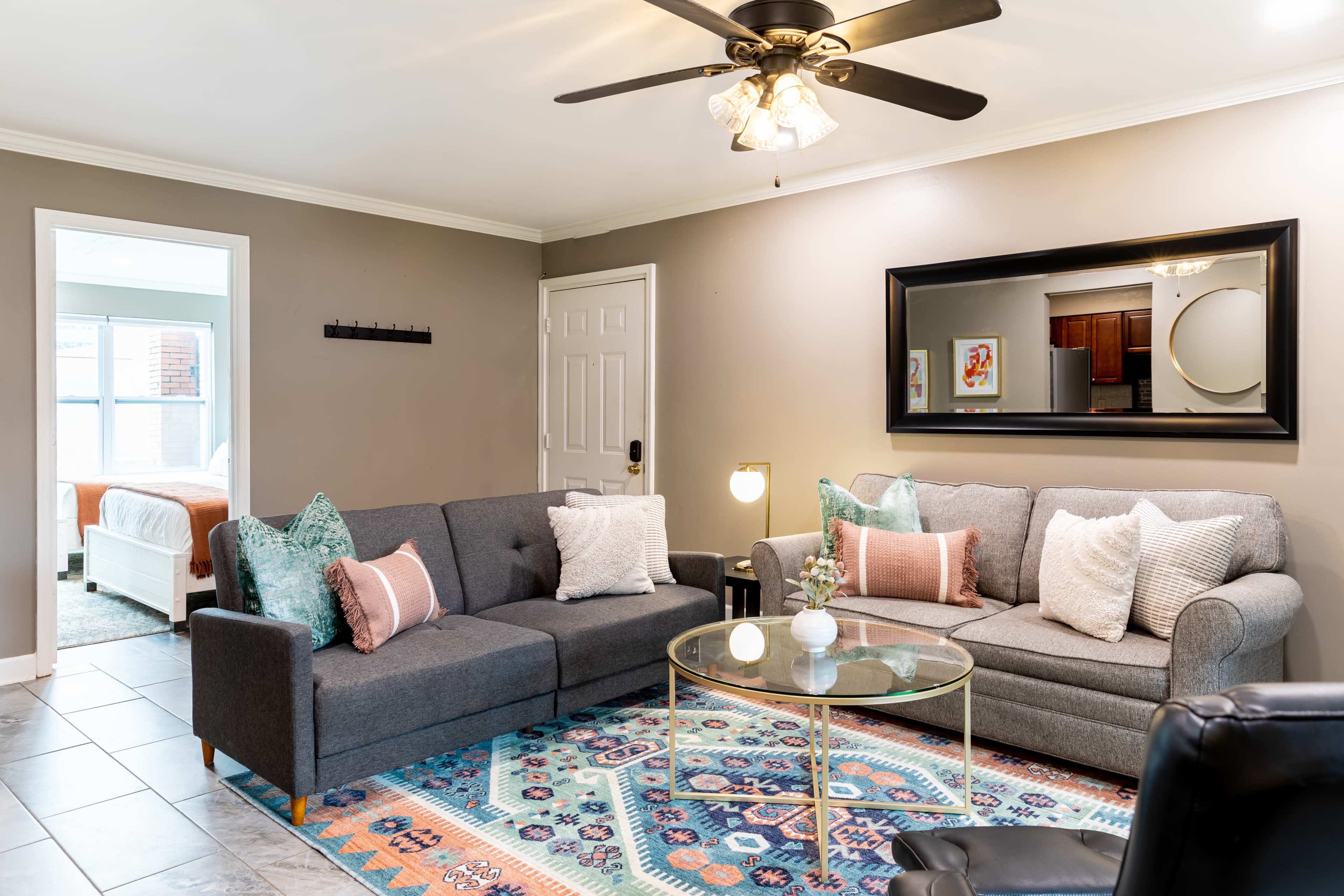 The living room features a gray sectional sofa and a gray couch, with a glass coffee table centered on a colorful area rug, and a doorway leading to a bedroom in the background.
