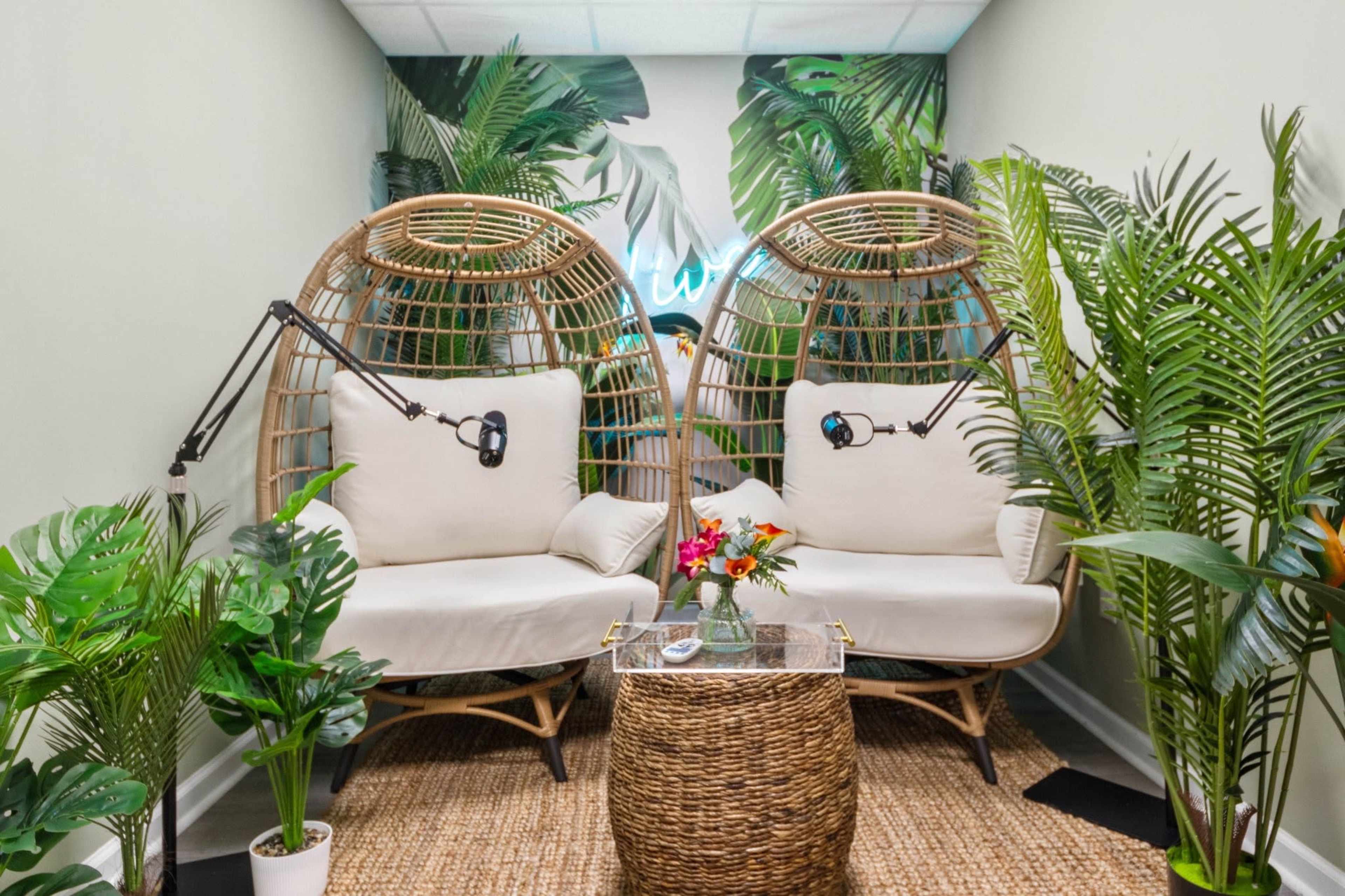 The image shows two rattan chairs positioned beside a small table, surrounded by greenery and tropical-themed decor.