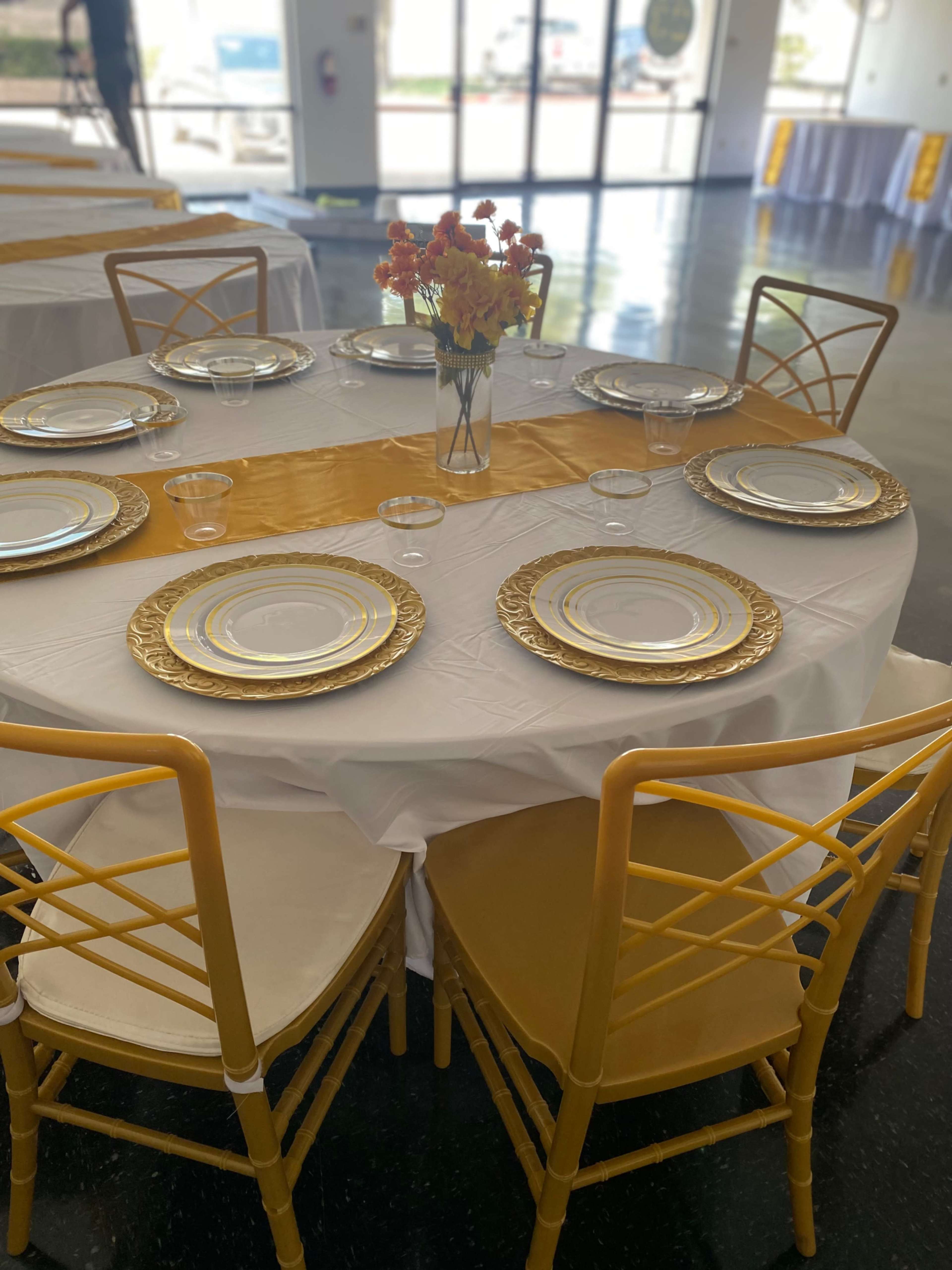 A round table is set with gold-trimmed plates, a yellow runner, clear glasses, and a vase of flowers, surrounded by yellow chairs.