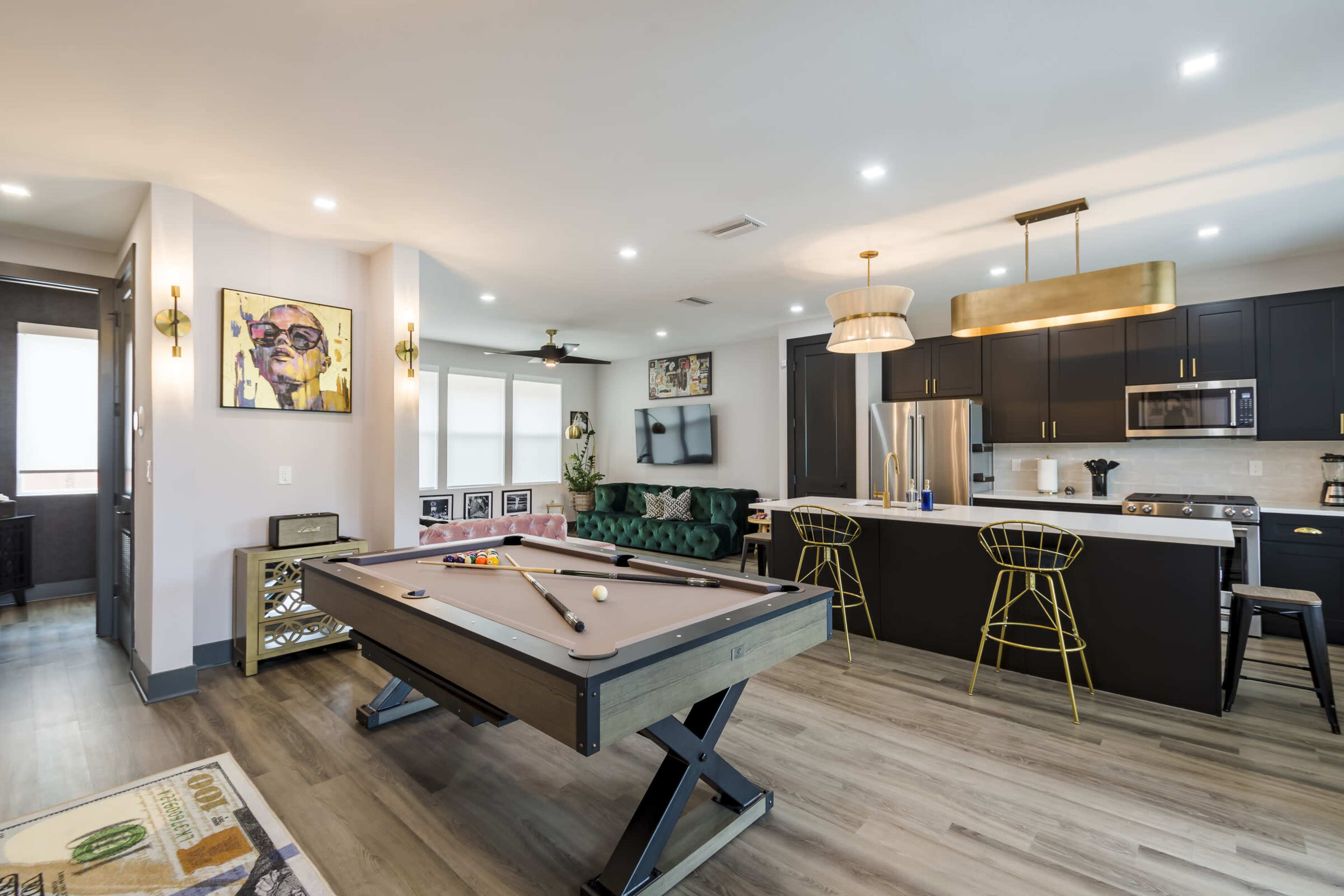 The image shows a modern living space featuring a billiards table in the center, surrounded by a stylish seating area and a kitchen bar with gold accents.