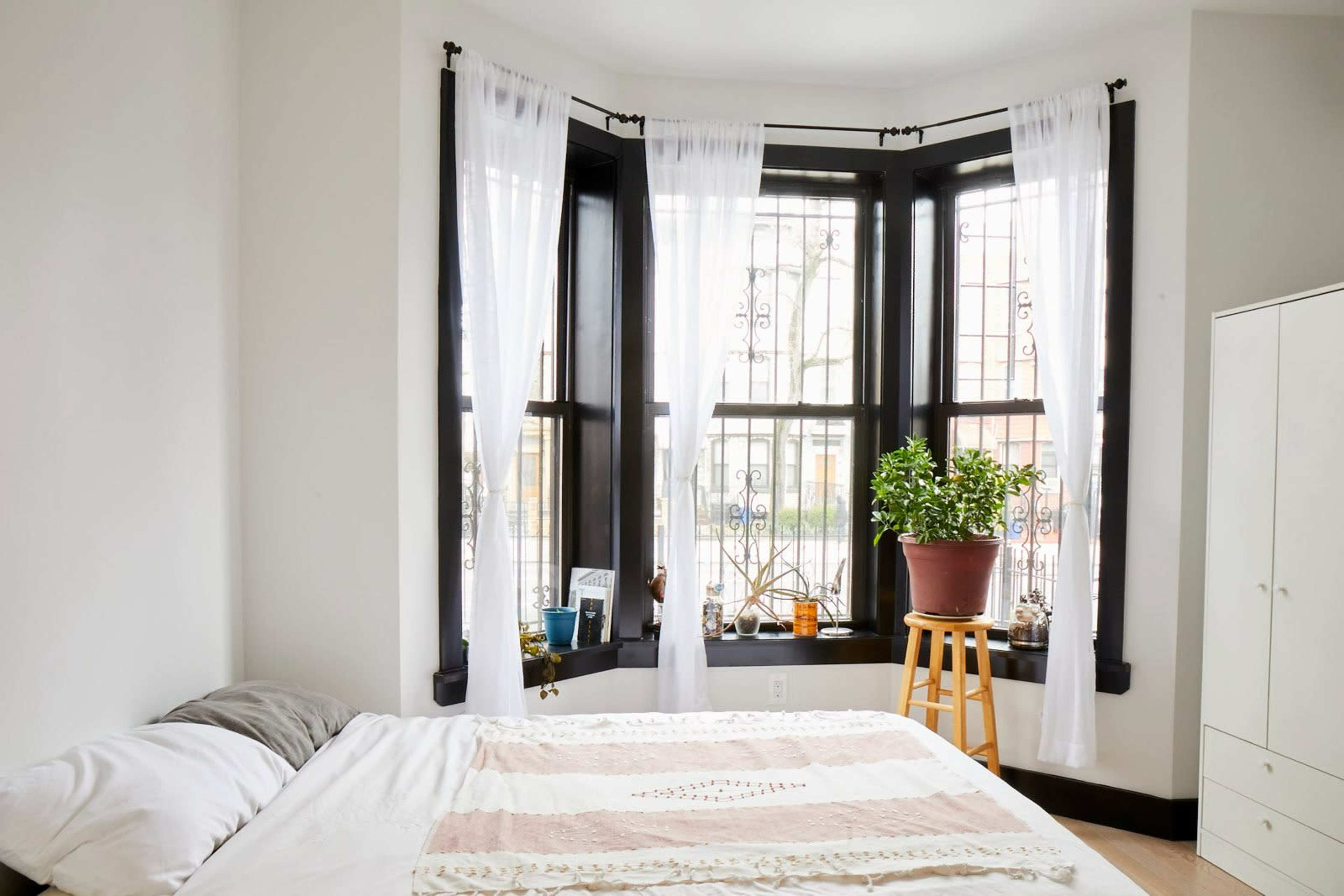 The image shows a bedroom with a bed positioned against a wall near a bay window adorned with sheer white curtains and a potted plant on a stool.