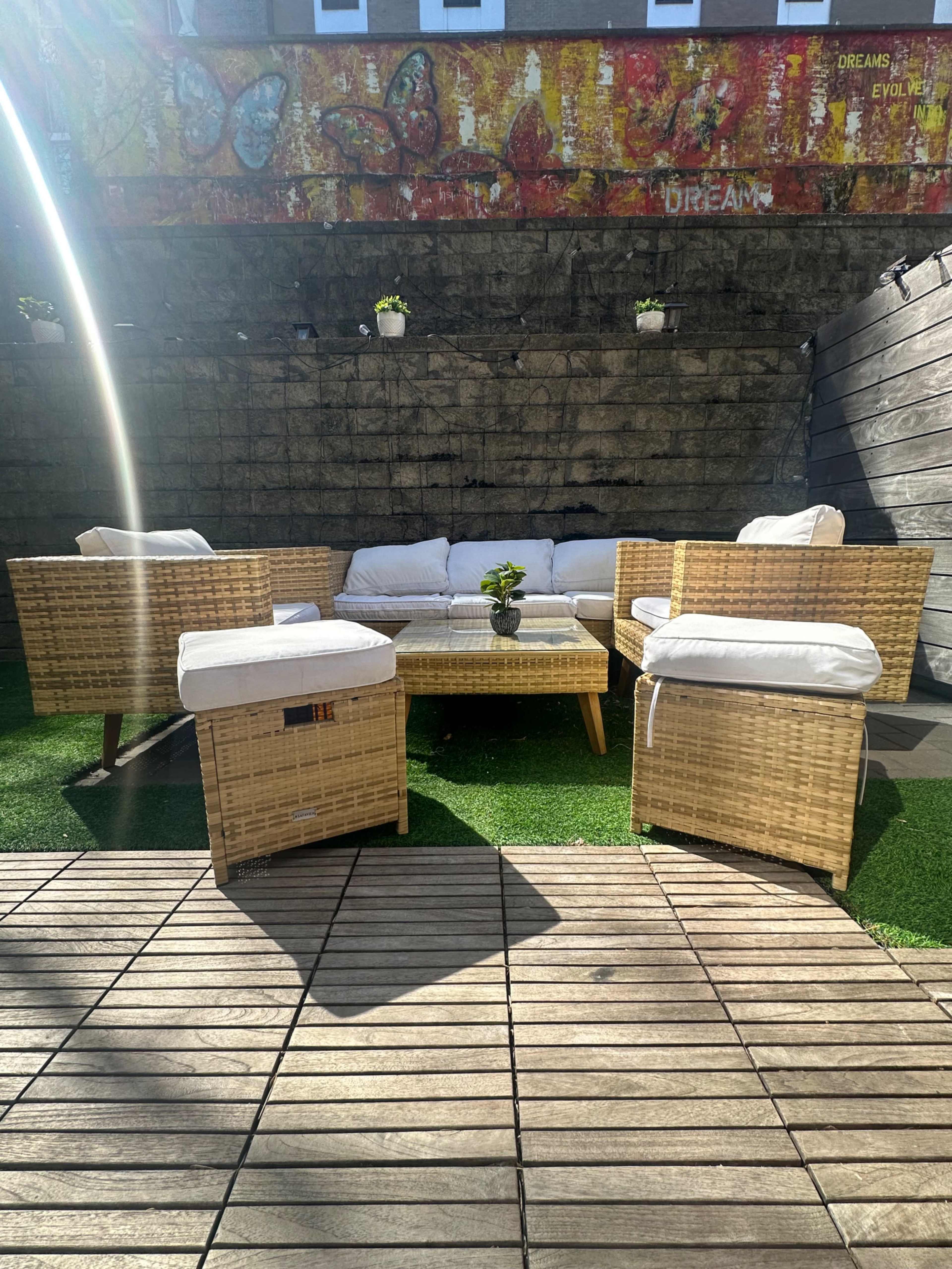 A seating area with wicker furniture is arranged on a wooden deck surrounded by artificial grass and a decorative wall featuring colorful butterfly art.