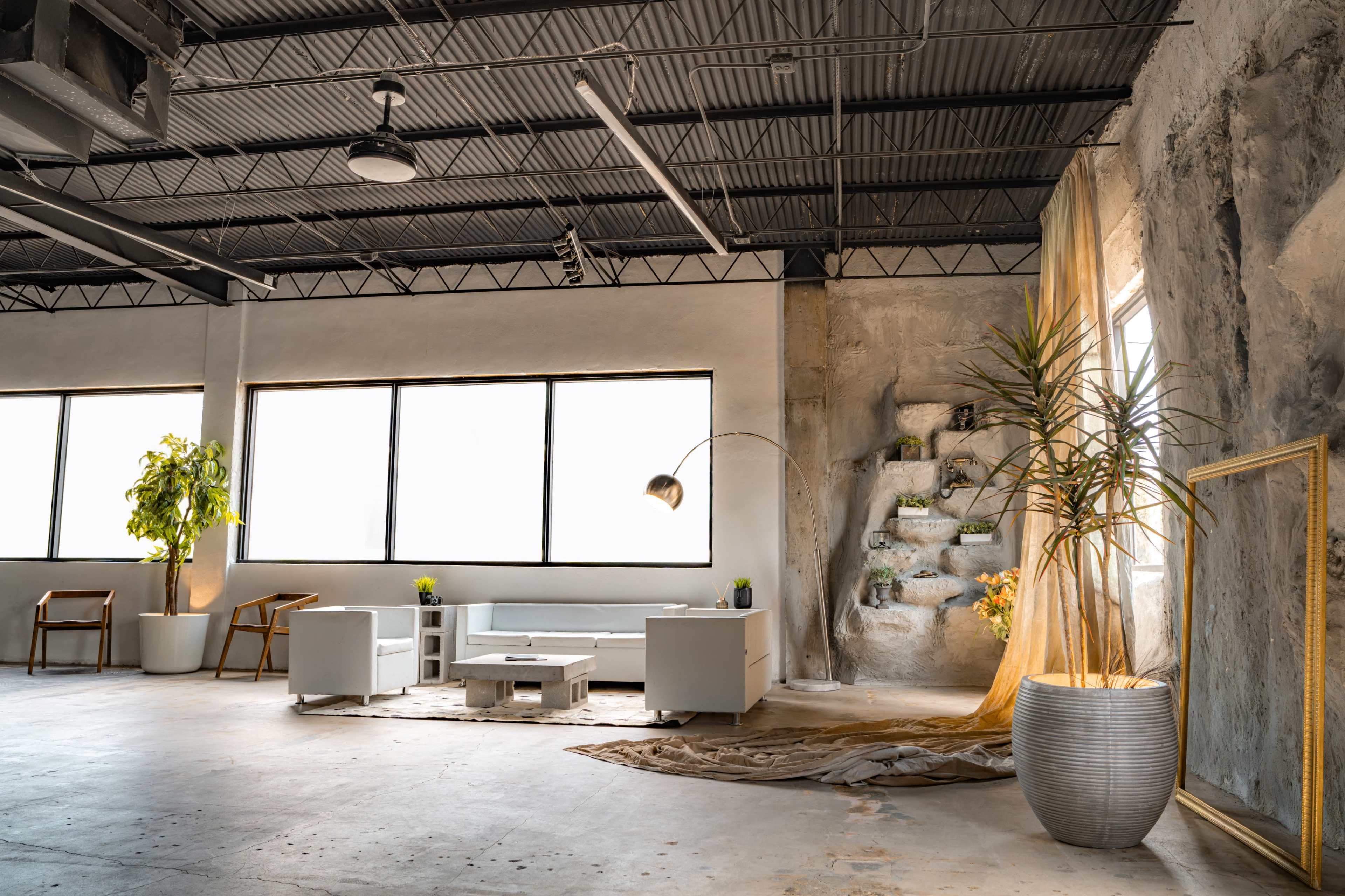 The scene features a modern, open space with a minimalist design, showcasing white furniture, large windows, and greenery against a concrete backdrop.