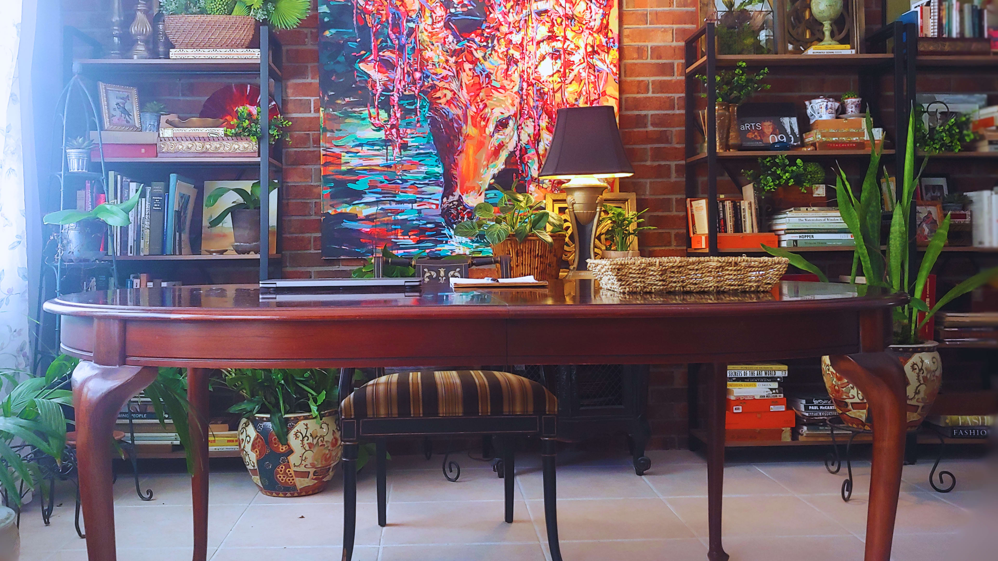A wooden desk is positioned in front of a colorful abstract painting, surrounded by plants and bookshelves filled with books and decorative items.