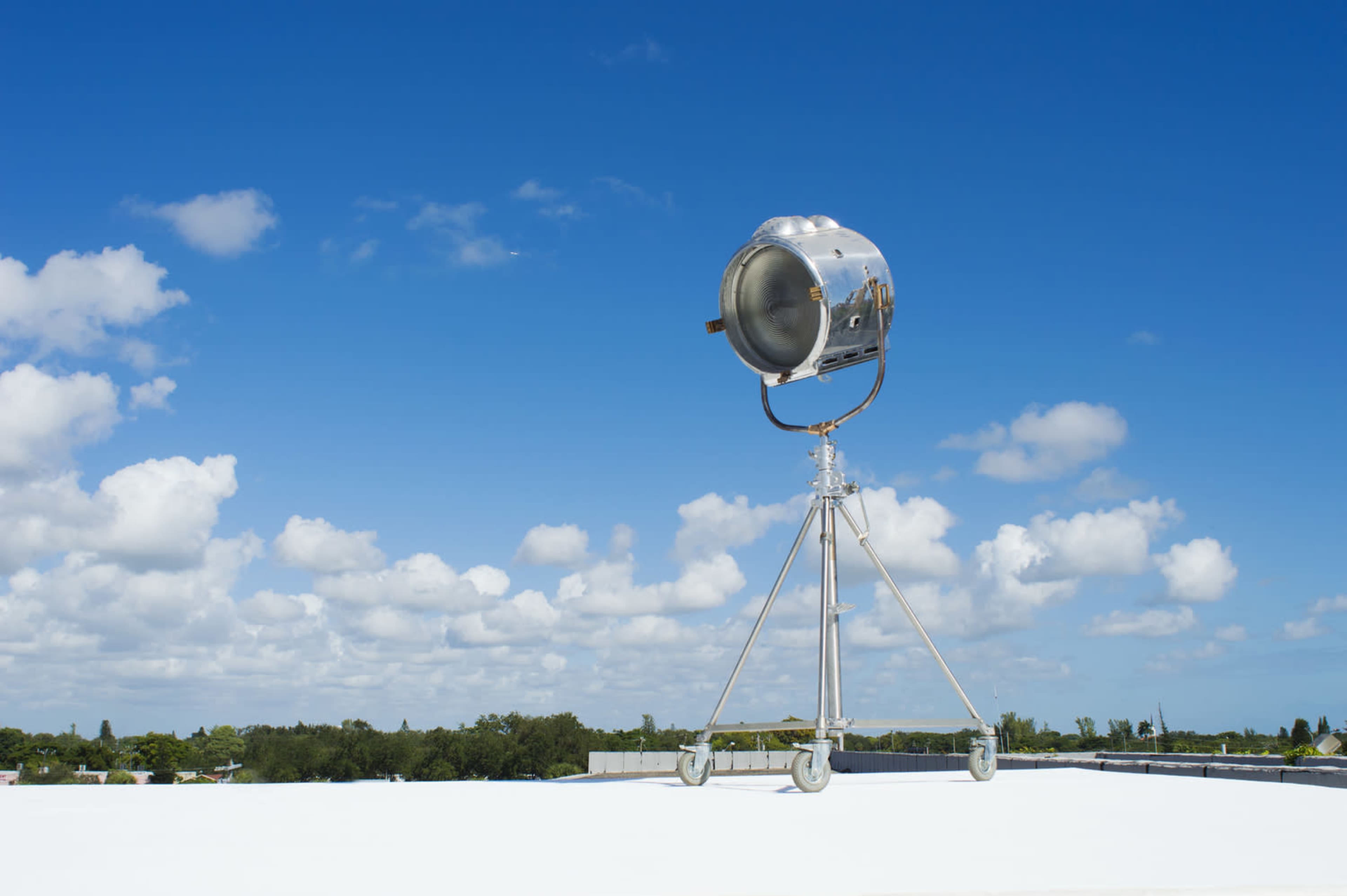 A large, chrome spotlight is mounted on a wheeled tripod on a flat rooftop under a blue sky with scattered clouds.