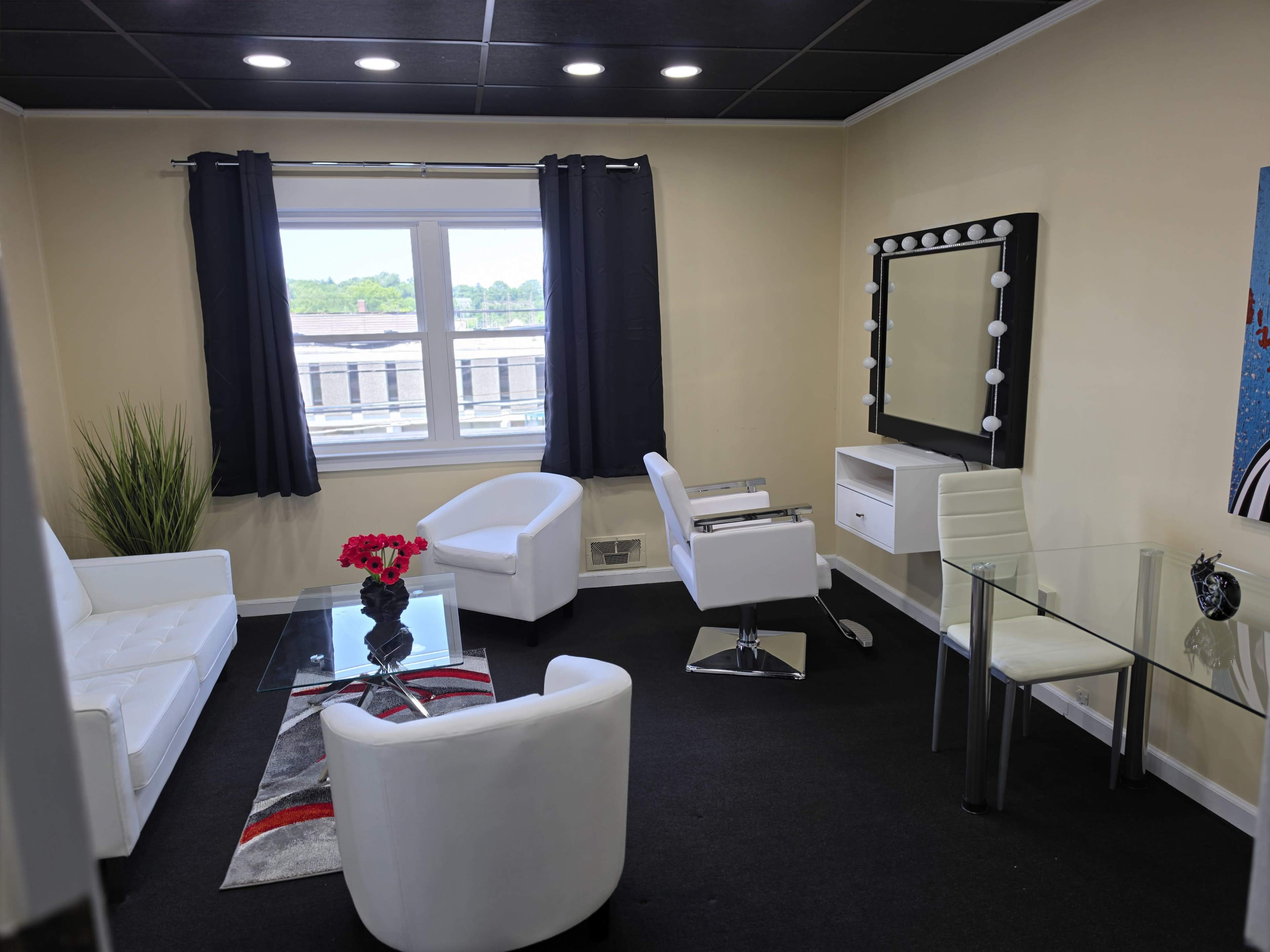The image shows a modern, well-lit room furnished with white sofas, a glass table, a mirror with lights, and decorative plants.
