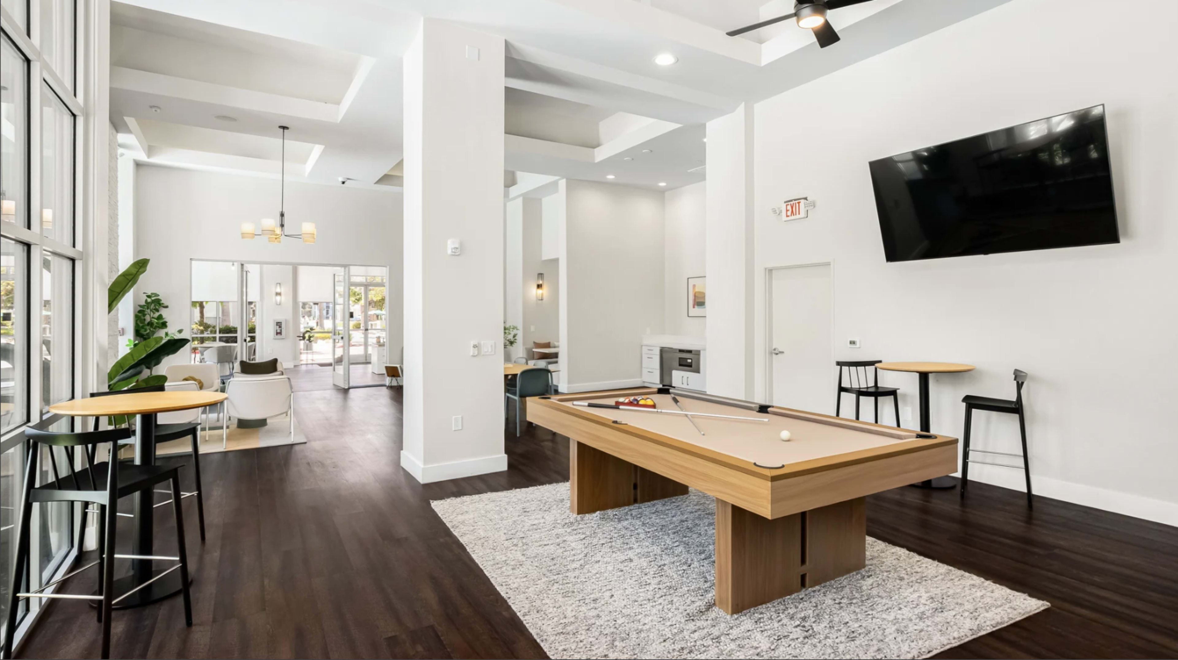 The image shows a modern lounge area featuring a pool table, a large television mounted on the wall, and dining tables with chairs.