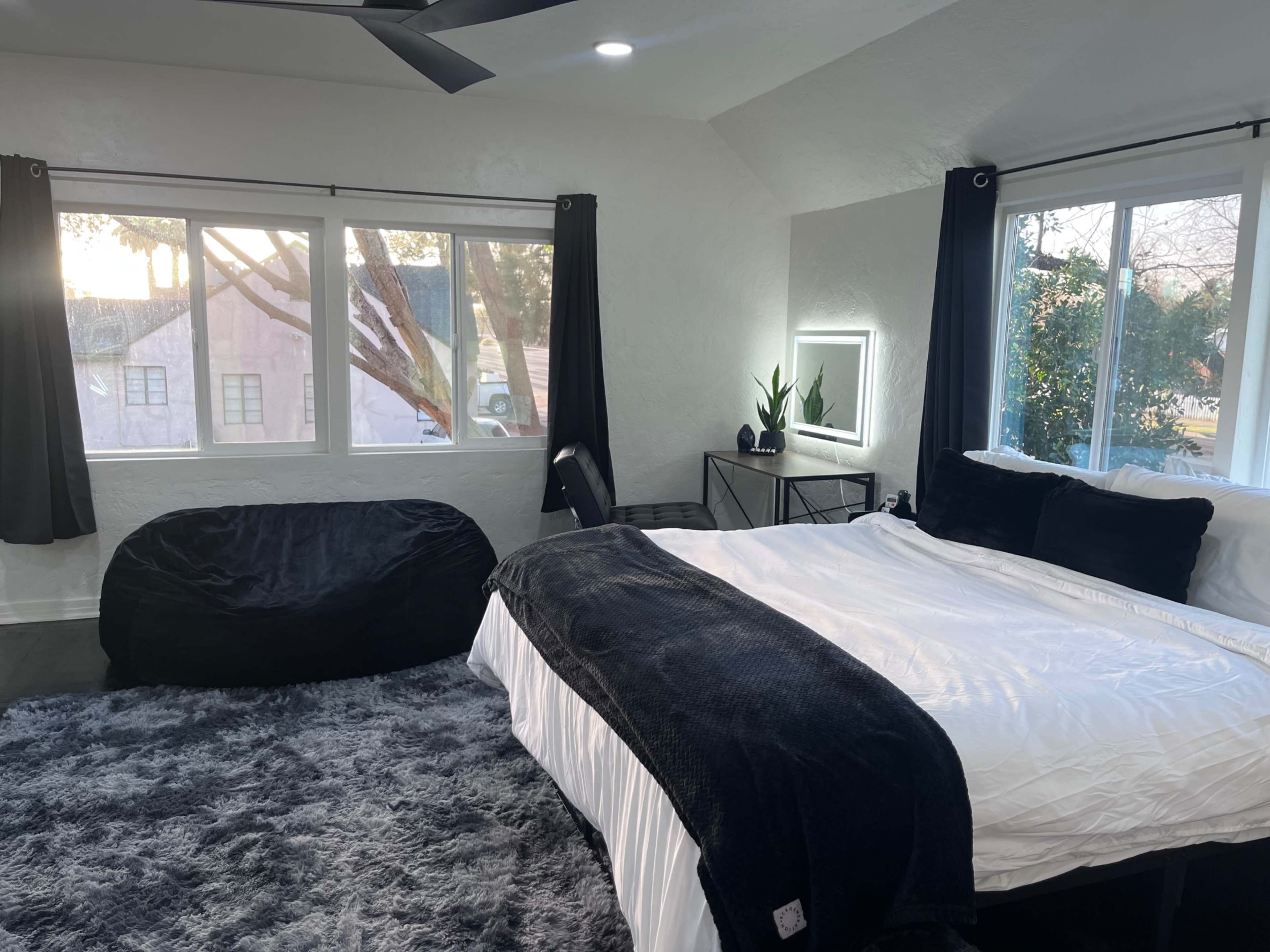 A bedroom features a large bed with black bedding, a bean bag chair, and a window with natural light filtering in.