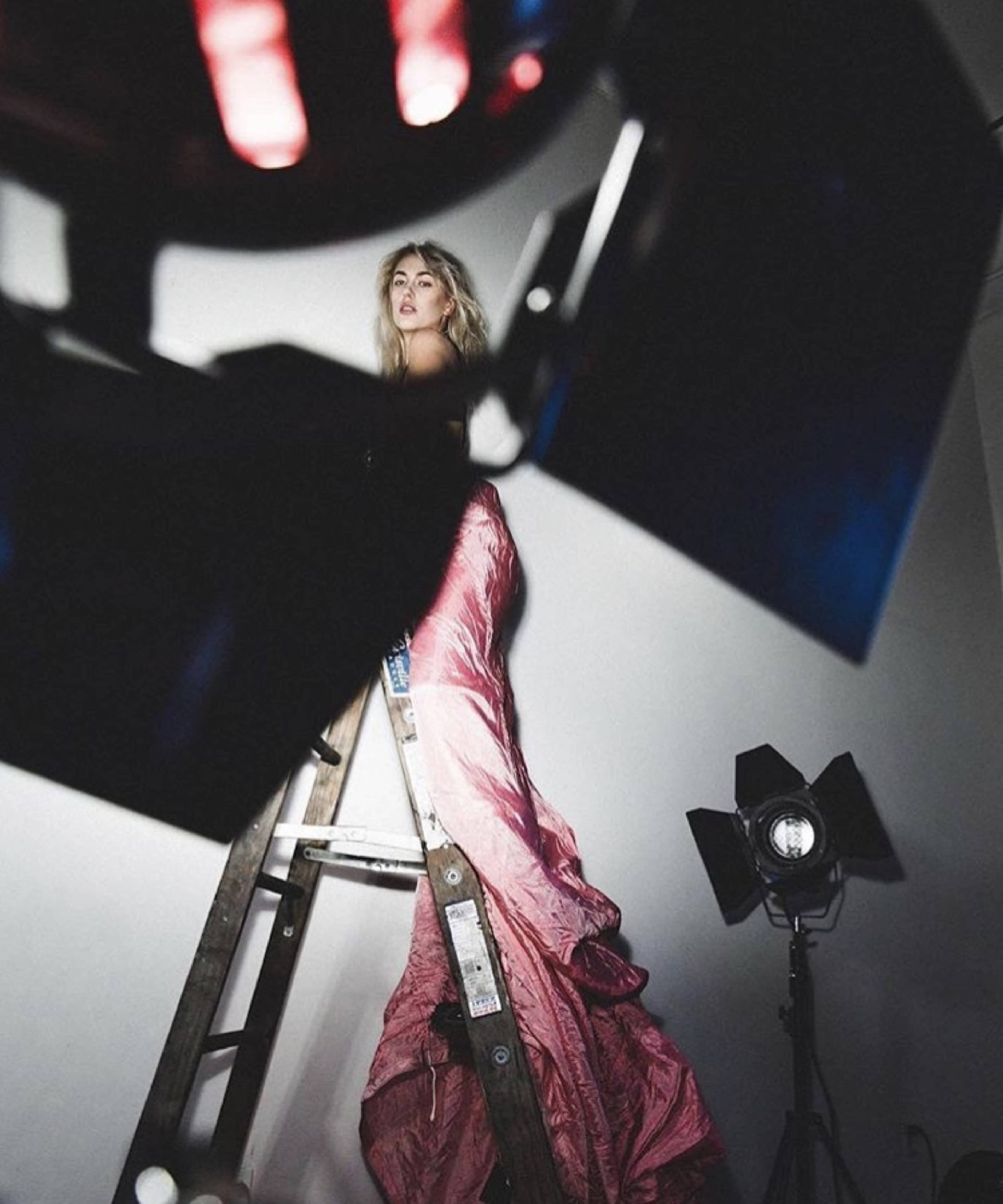 A model poses on a ladder while wearing a long, flowing garment, with studio lights illuminating the scene.