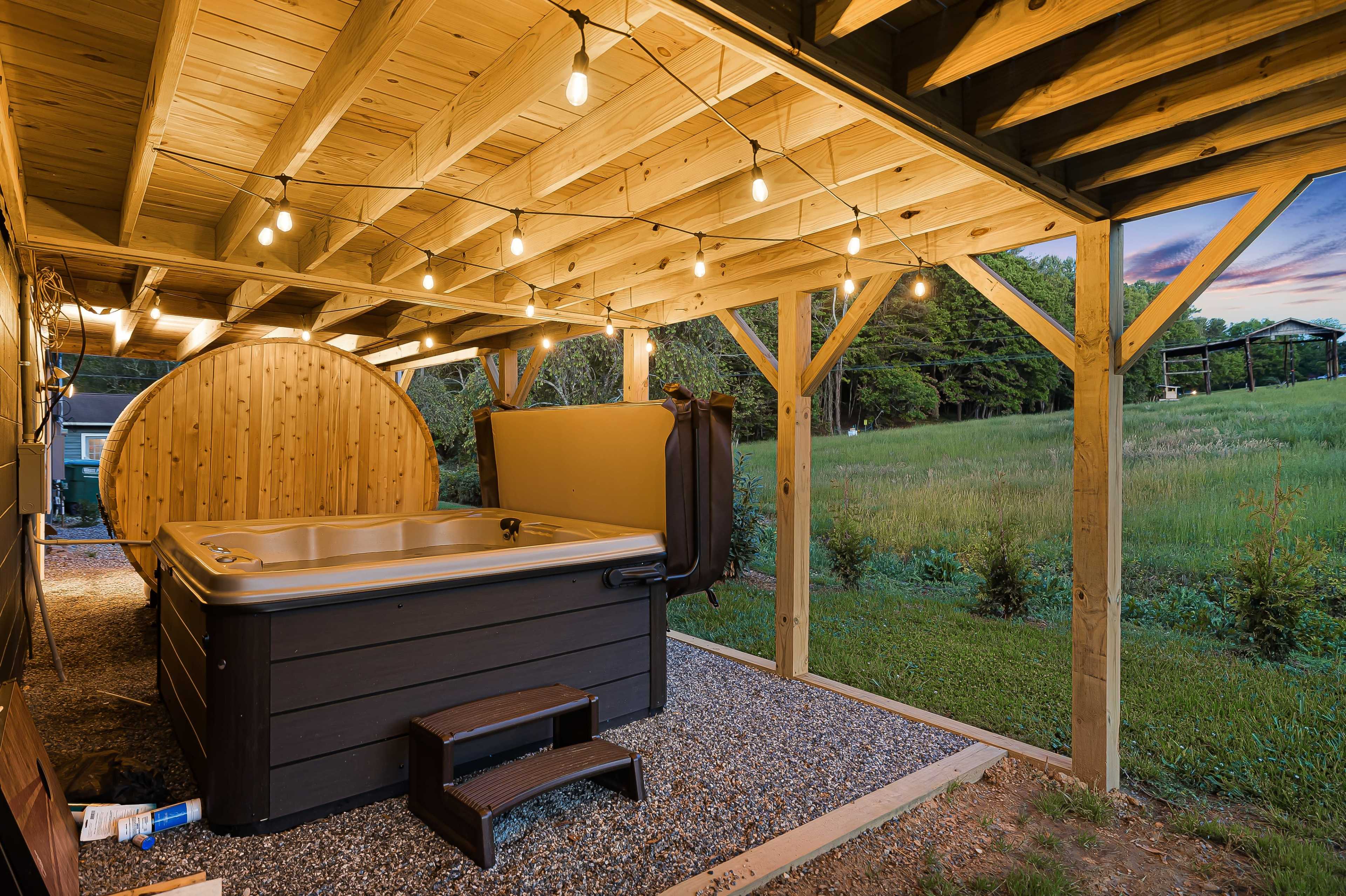 The image shows a wooden terrace with a hot tub and a barrel-shaped sauna, all illuminated by string lights, surrounded by a grassy landscape.