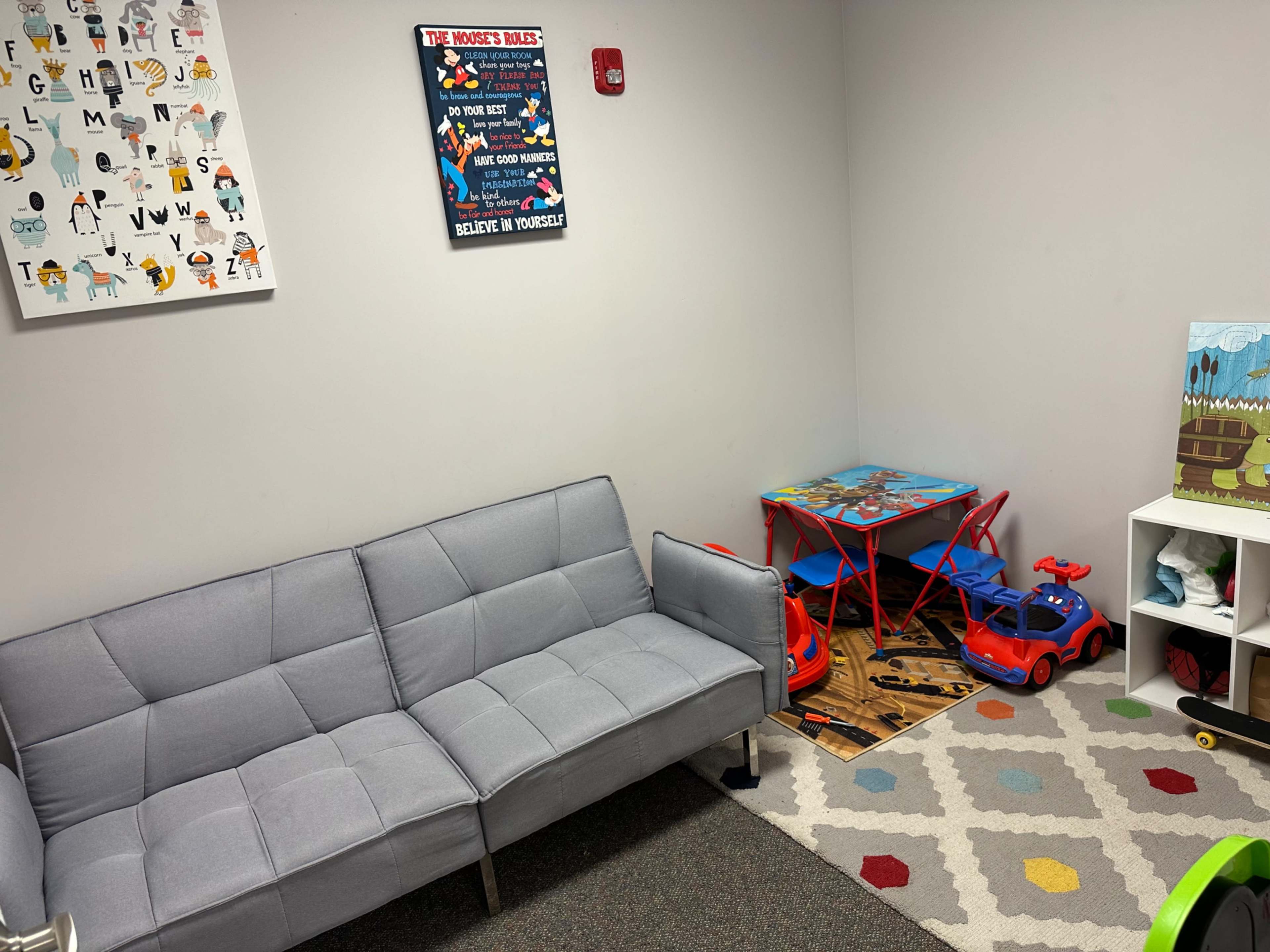 A small room featuring a gray sofa, a colorful area rug, children's toys, and a table with chairs in the corner.