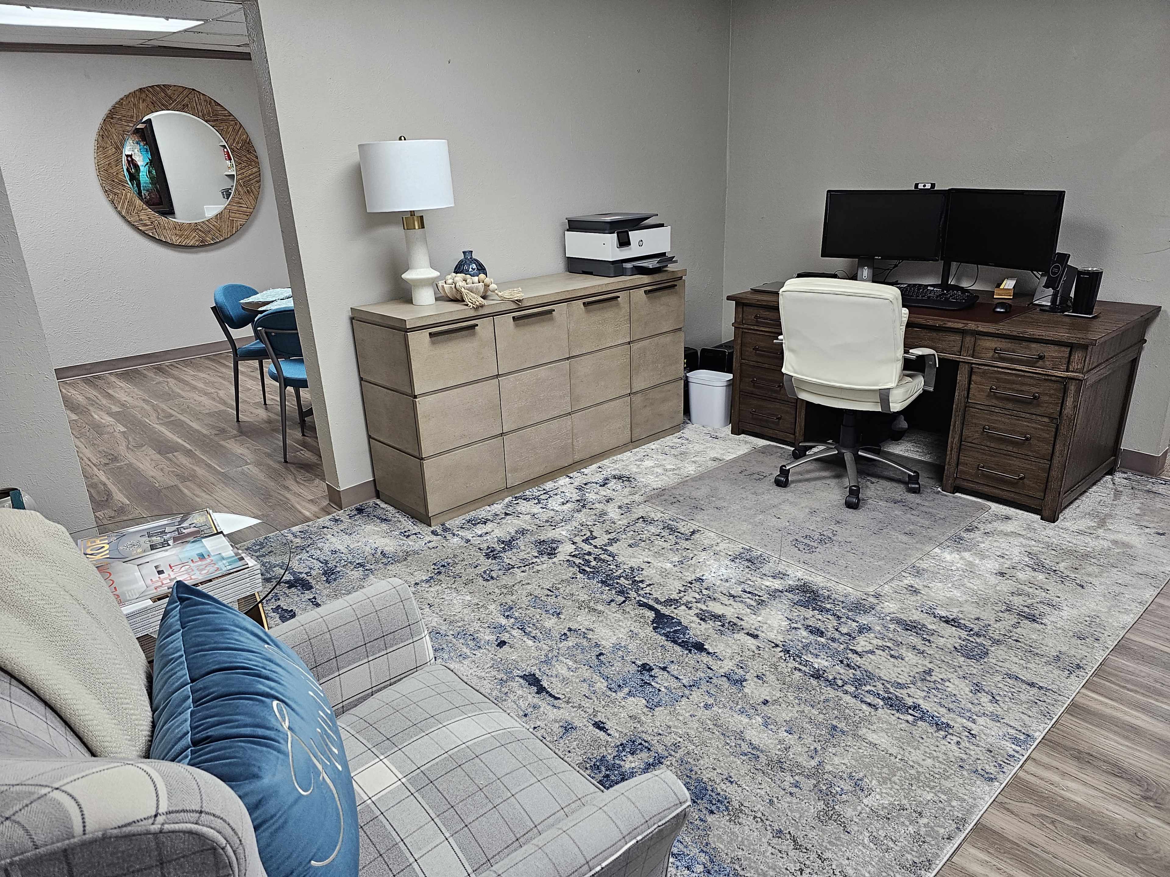 A small office space features a wooden desk with a computer, a printer, a light-colored sofa, a dresser, and a round mirror on the wall.