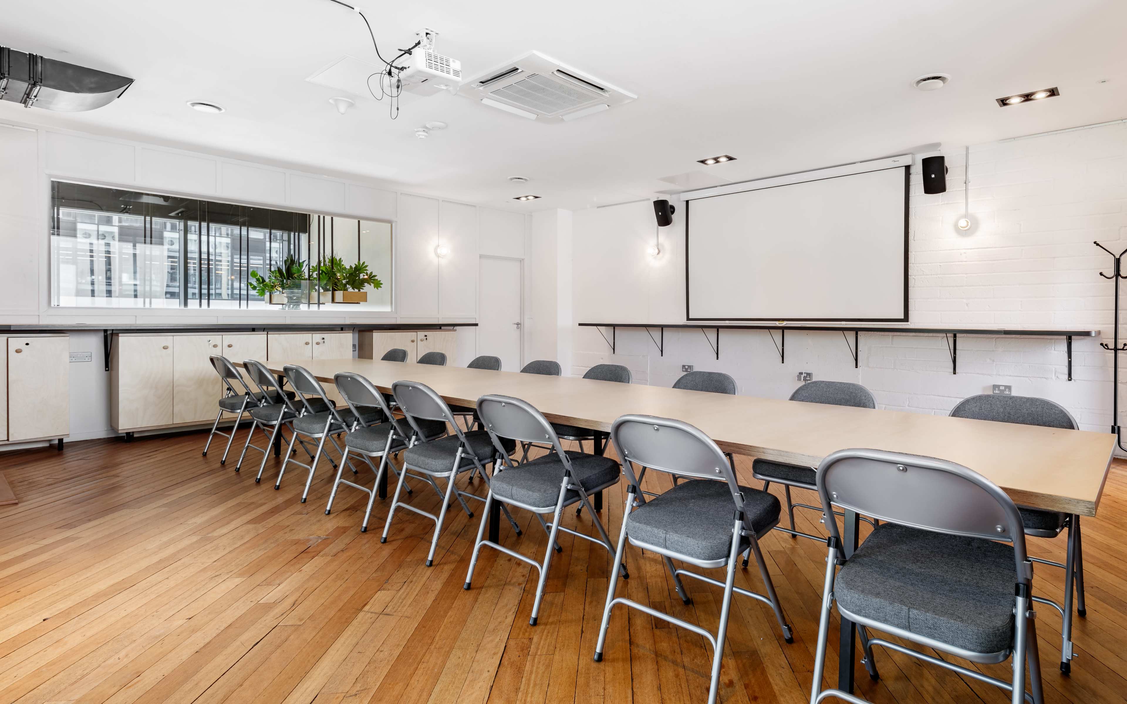 A long wooden table is surrounded by metal chairs in a well-lit conference room with a large screen and a mirrored wall.