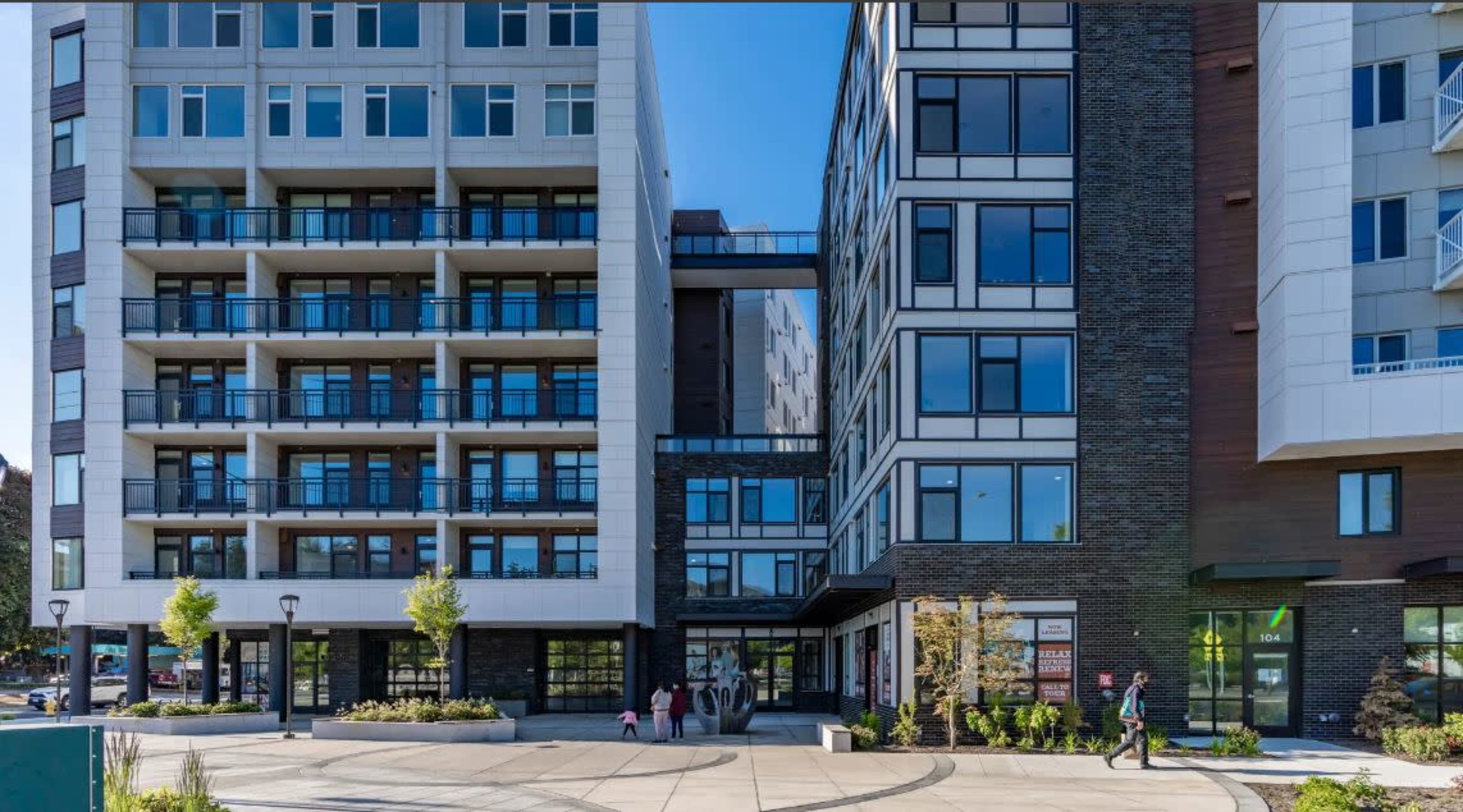 A multi-story modern building features a mix of glass and brick facades, with outdoor seating areas and pedestrians in the foreground.