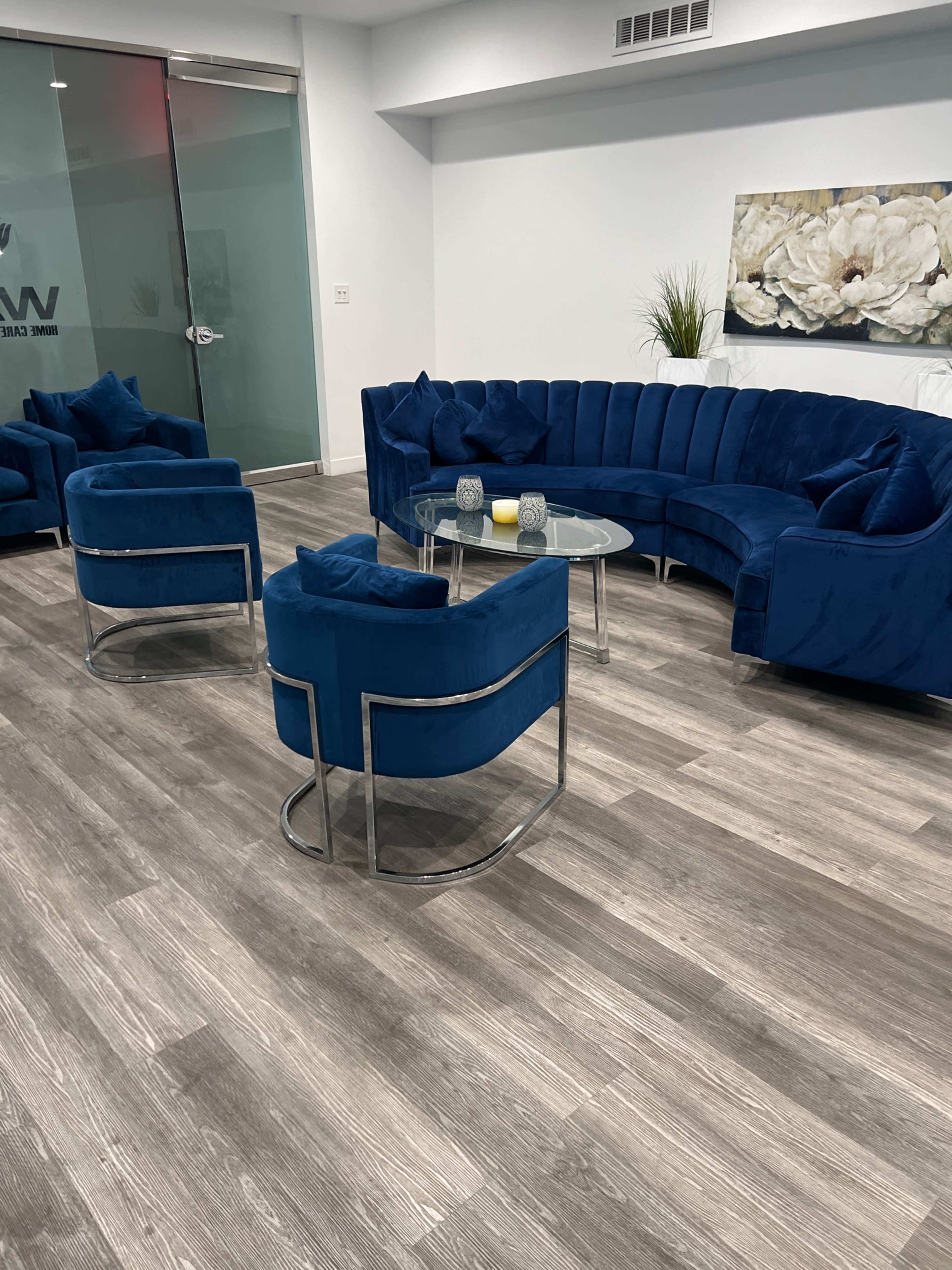 A modern waiting area with a curved blue sectional sofa and matching armchairs arranged around a glass coffee table on grey wooden flooring.