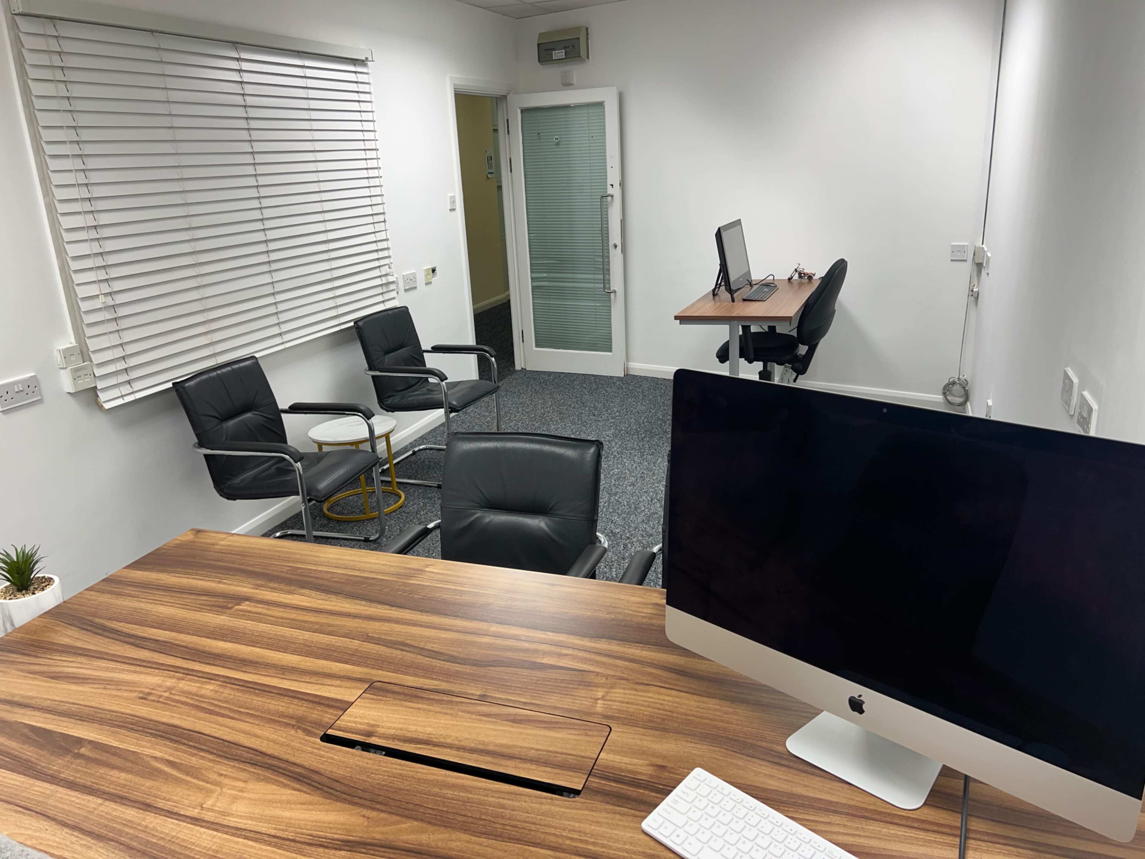 The image shows a modern office space with a wooden desk, an iMac computer, two black chairs, and a separate desk in the background.