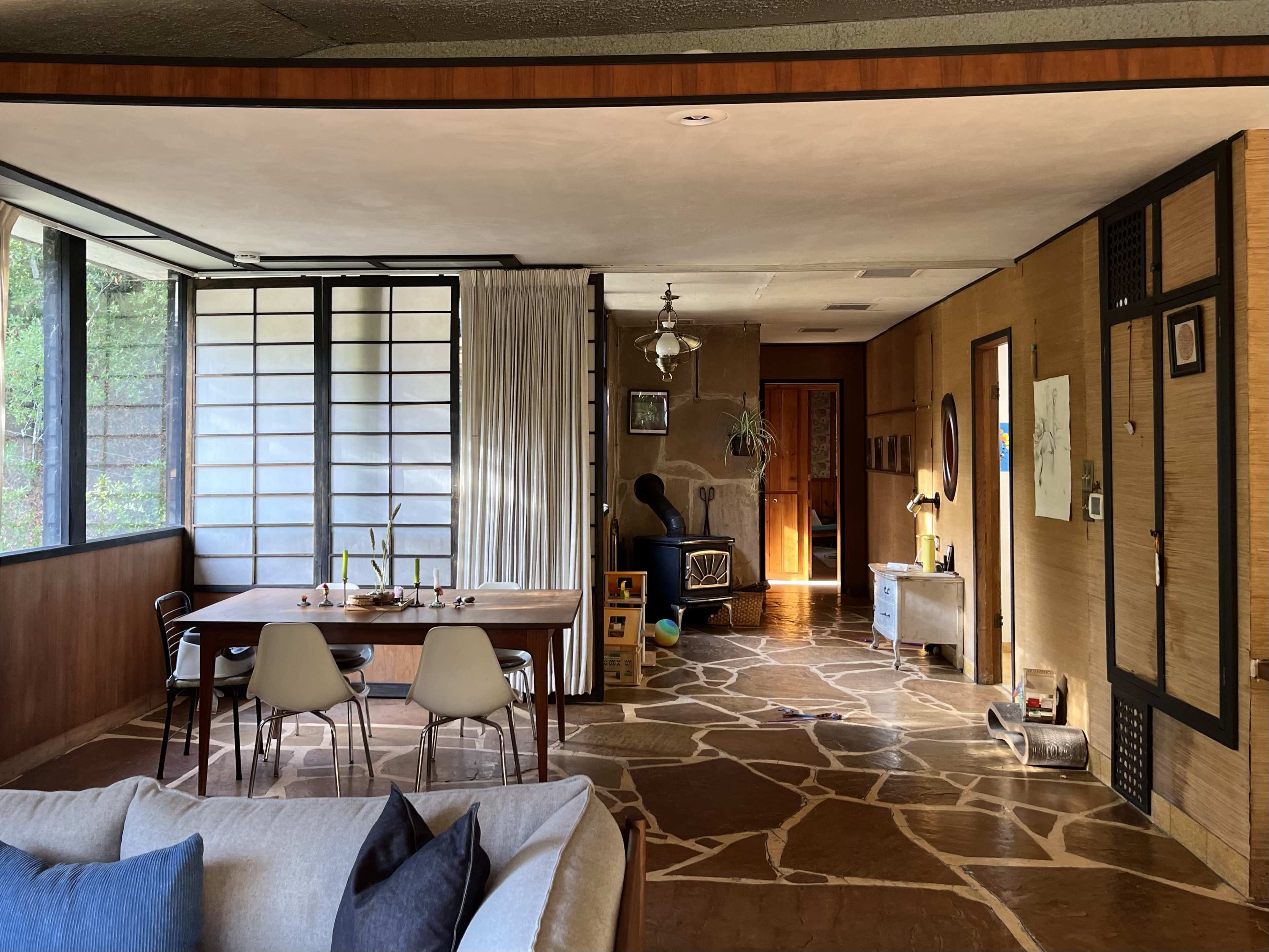 The image shows a spacious, modern living area with a dining table, chairs, and a mix of natural light and shadow from large windows and an adjacent room.
