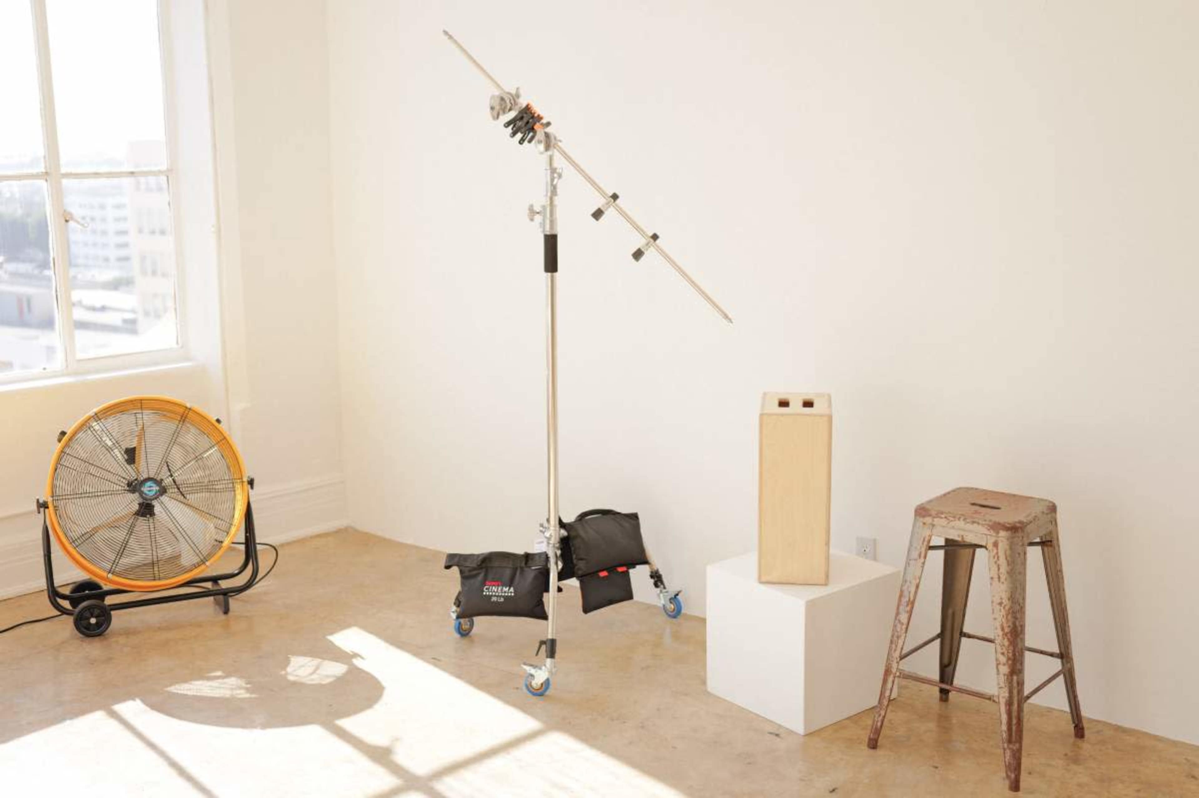 The image features a studio space with a large fan, a lighting stand with equipment, a wooden box, and a metal stool arranged in a bright room with natural light.