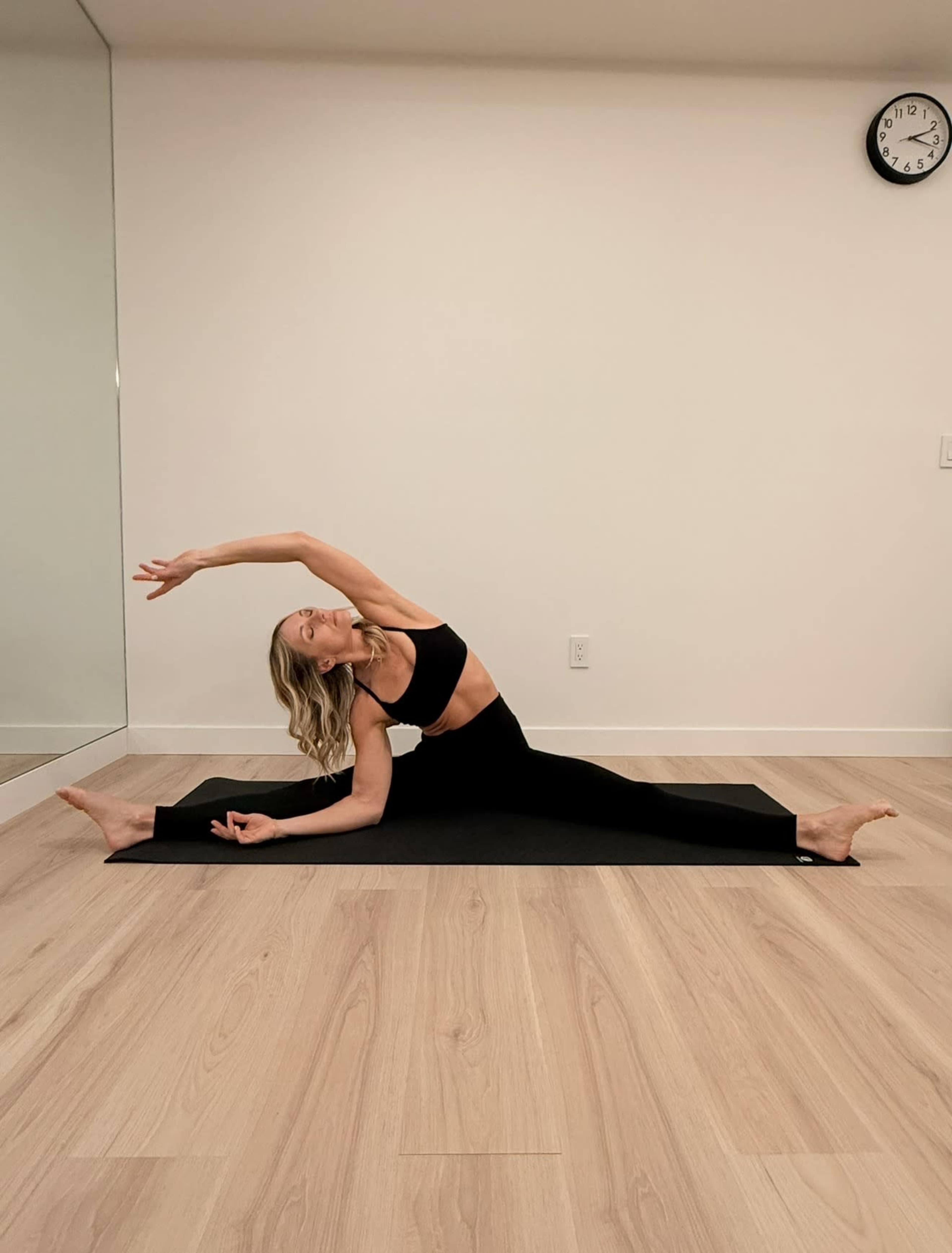 A woman in fitness attire performs a side stretch on a yoga mat in a minimalist room with a large mirror and a clock on the wall.