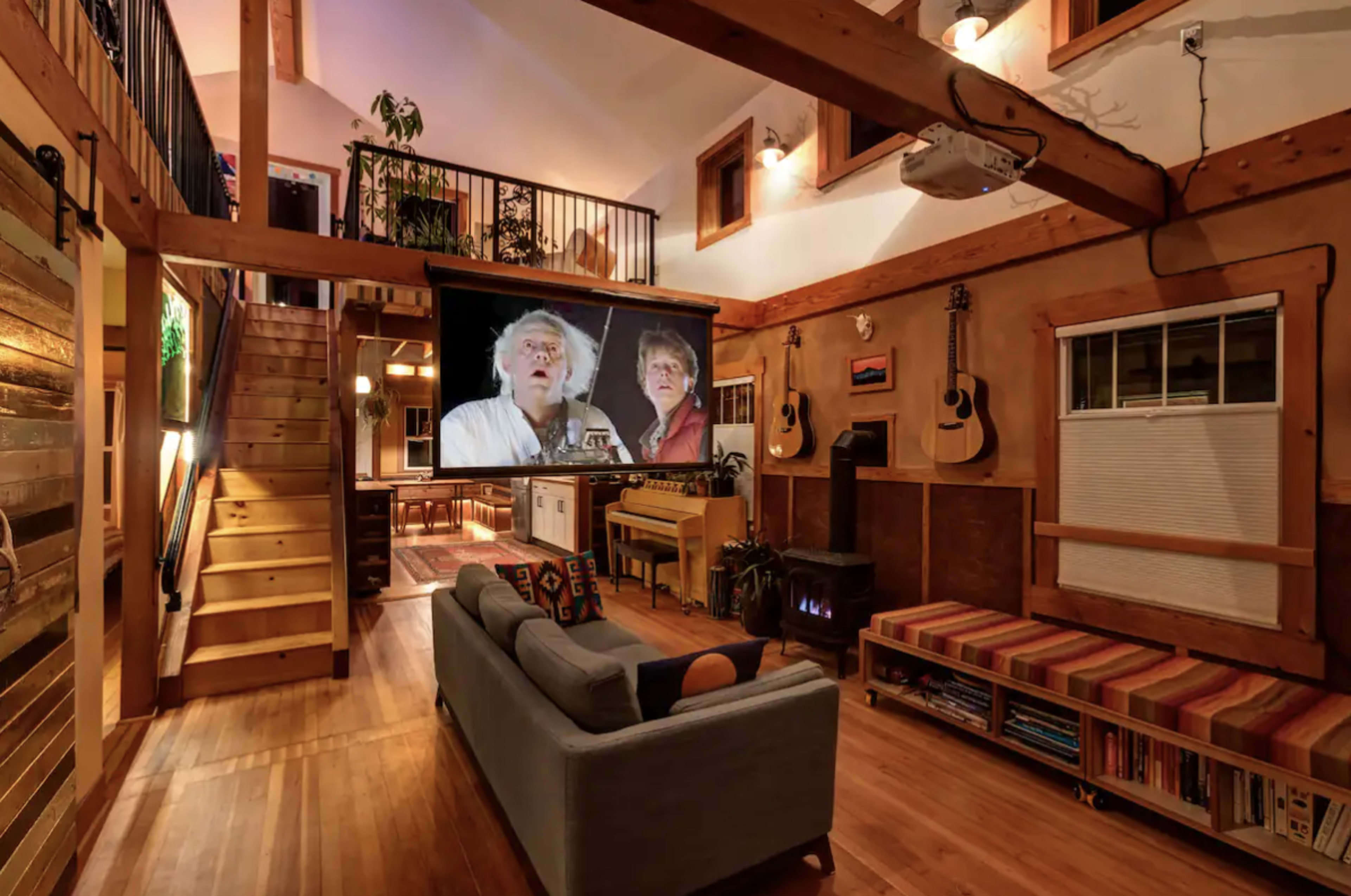 A spacious, wooden interior features a cozy seating area in front of a large screen displaying two people, with a staircase, musical instruments, and a fireplace in the background.
