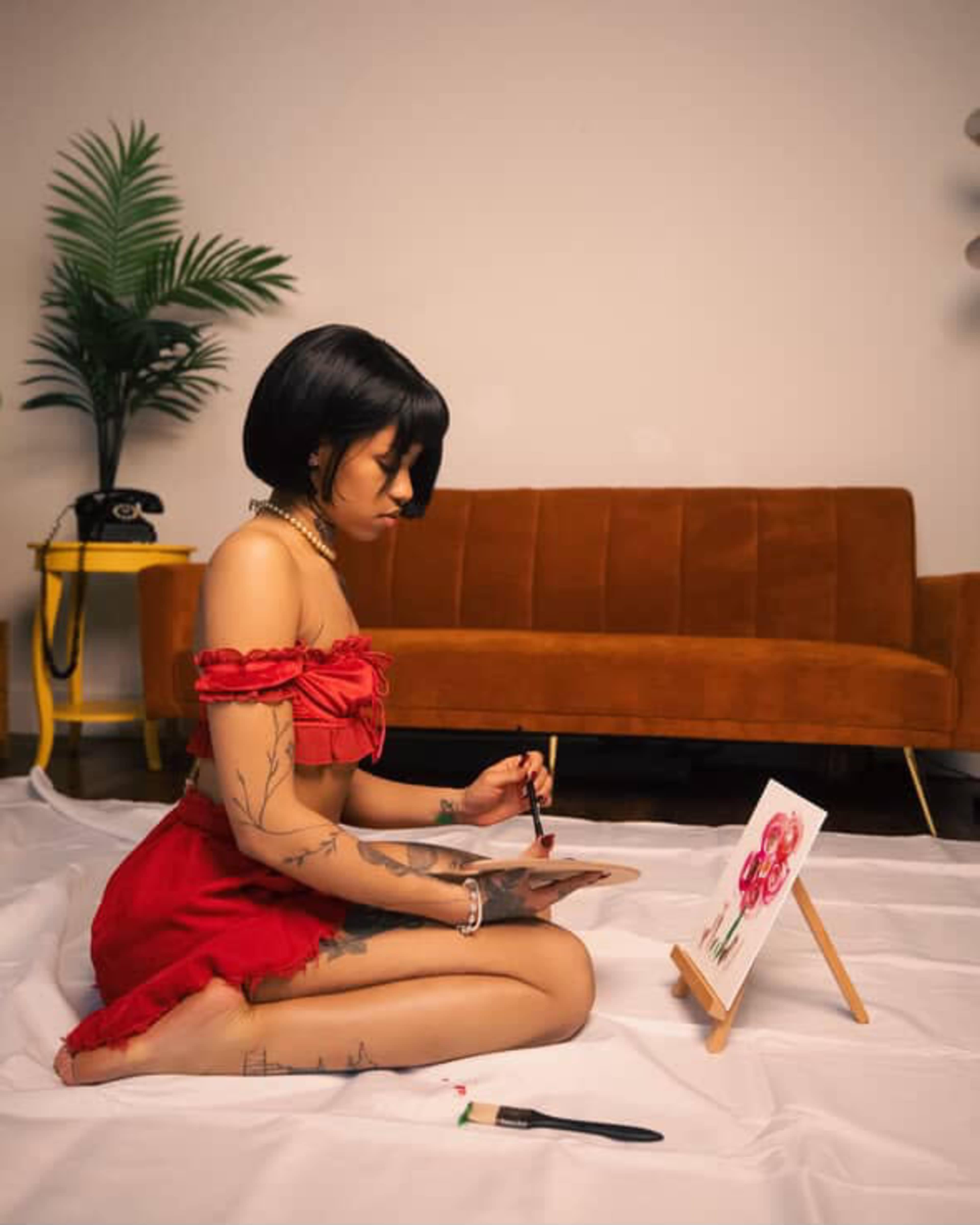 A woman with tattoos sits on the floor in a red outfit, painting flowers on a canvas while a brown couch and a plant are in the background.
