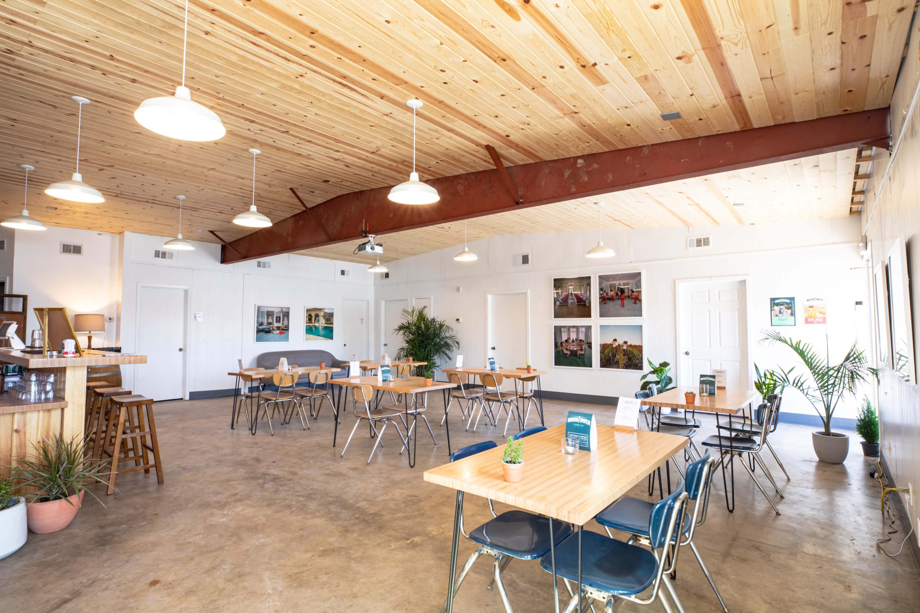 The image shows a spacious, minimalist interior of a café with wooden ceiling beams, metal chairs, and neatly arranged tables, along with potted plants and wall art.